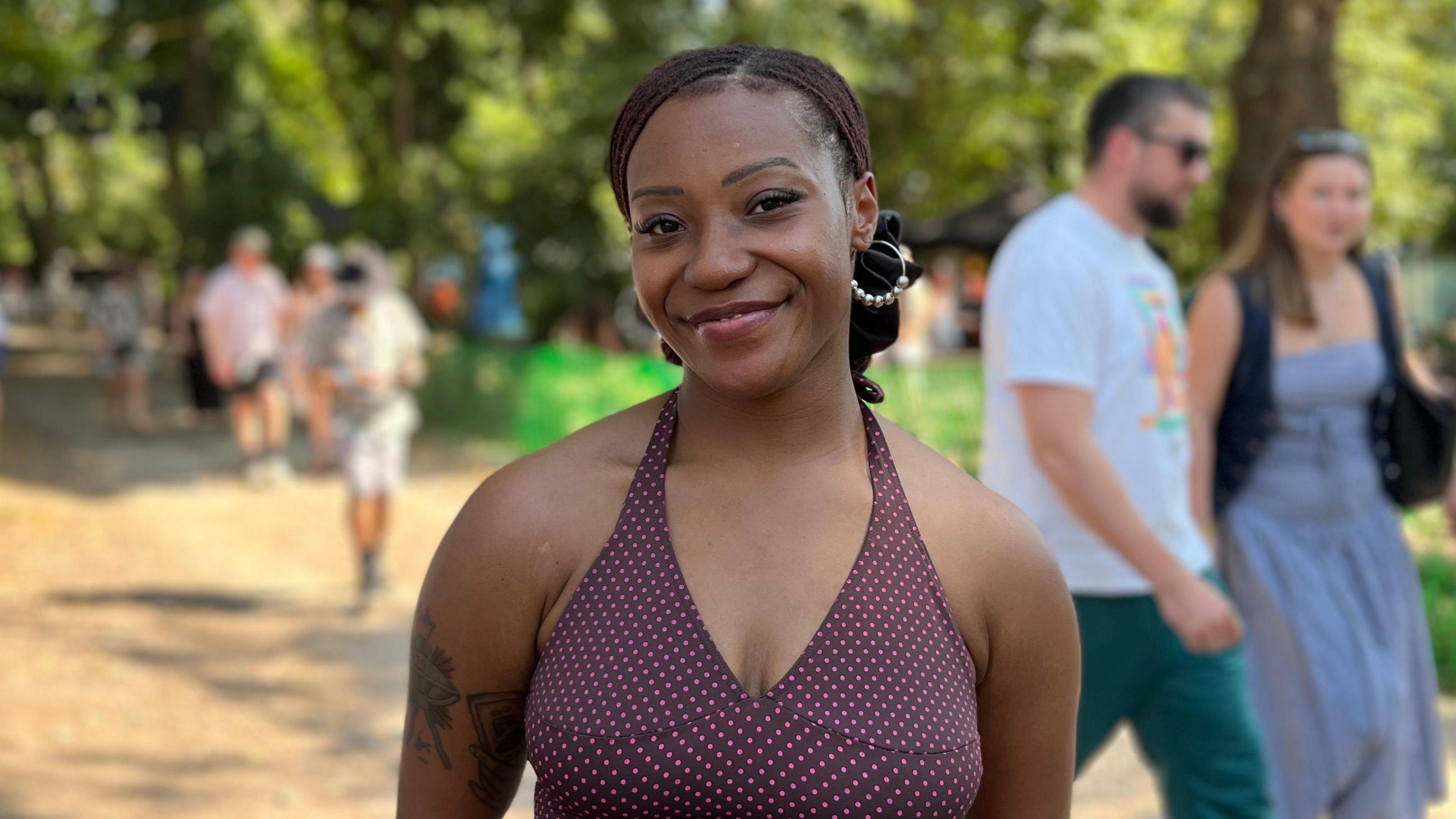 A woman is wearing a purple and pink spotted halter neck top and has her braided hair tied back in a scrunchie. She is standing on a festival site with trees as people walk around the grounds.