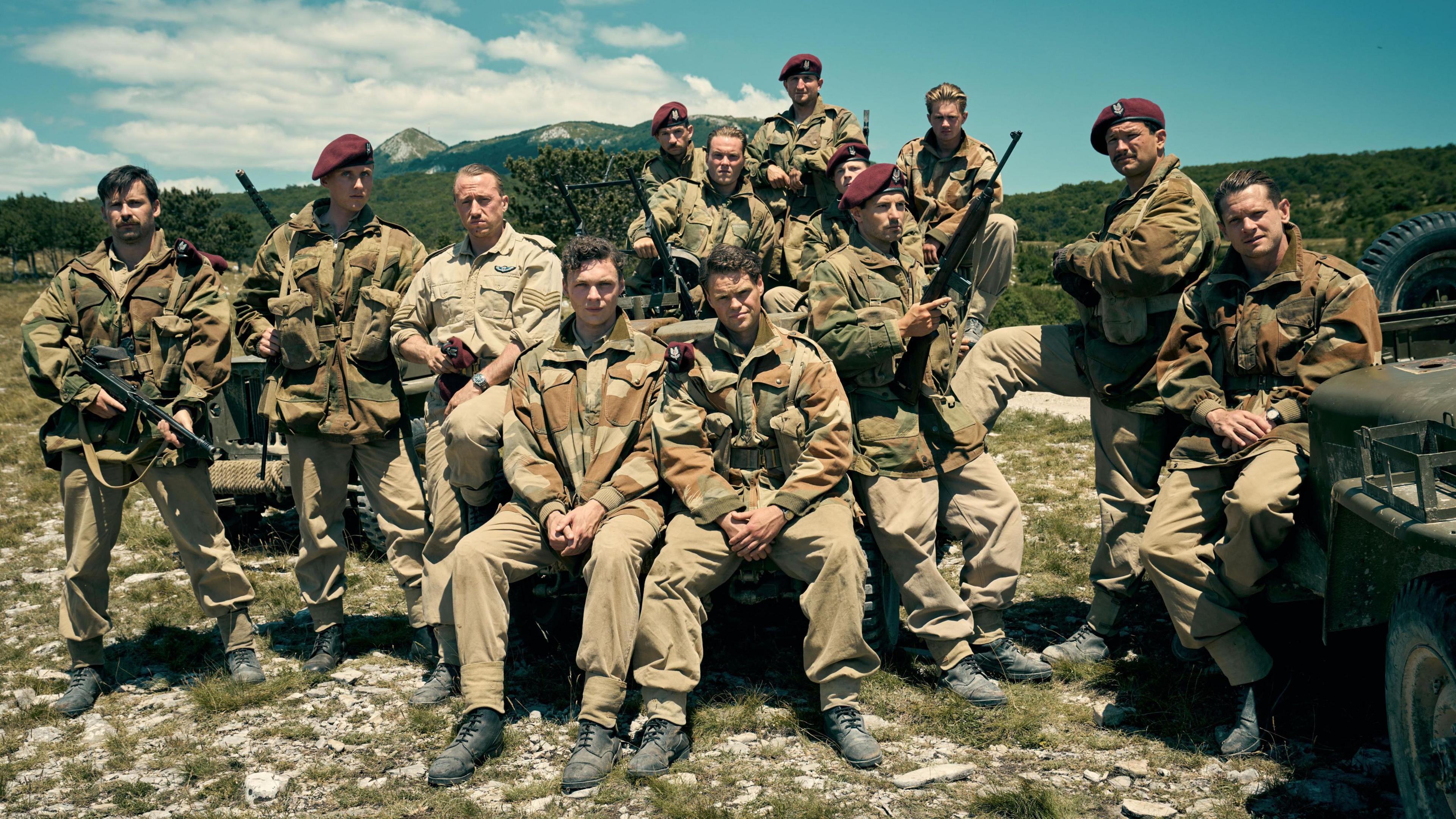 A photograph of the cast of SAS Rogue Heroes. Thirteen men in army uniform and purple berets. They are posing with guns on military vehicles and in a mountain setting.