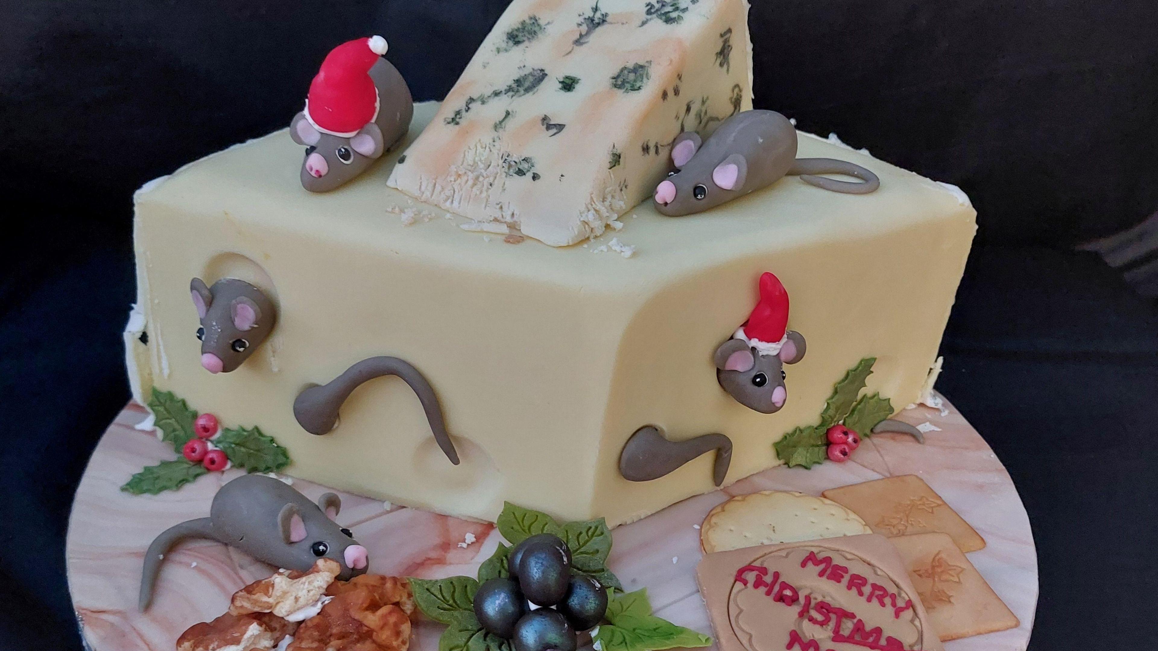 A cake that looks like a block of cheddar with mice poking out of it, on top of it is a block of stilton and two mice are on top of the cake, some of them wearing red Father Christmas hats