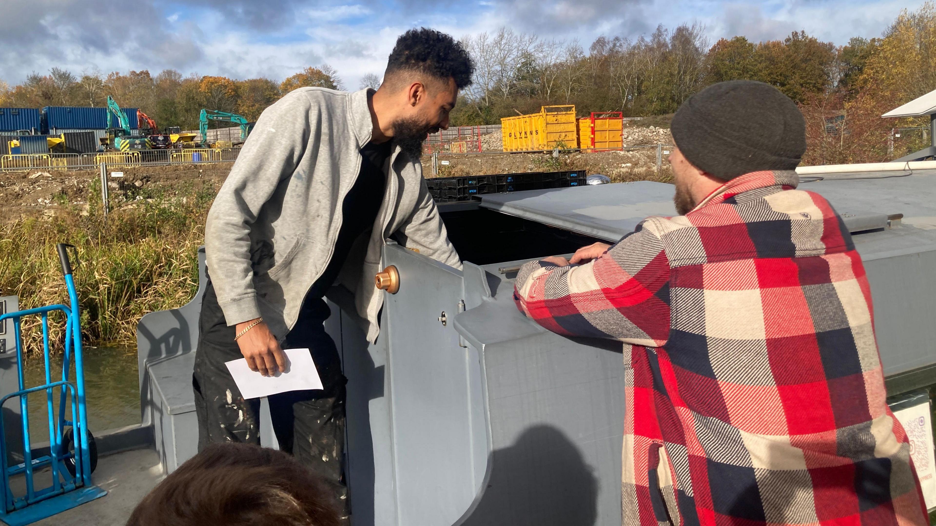 A man is dropping an unseen object into a hatch on his boat while Andy Simpson stands with his back to camera, talking to him