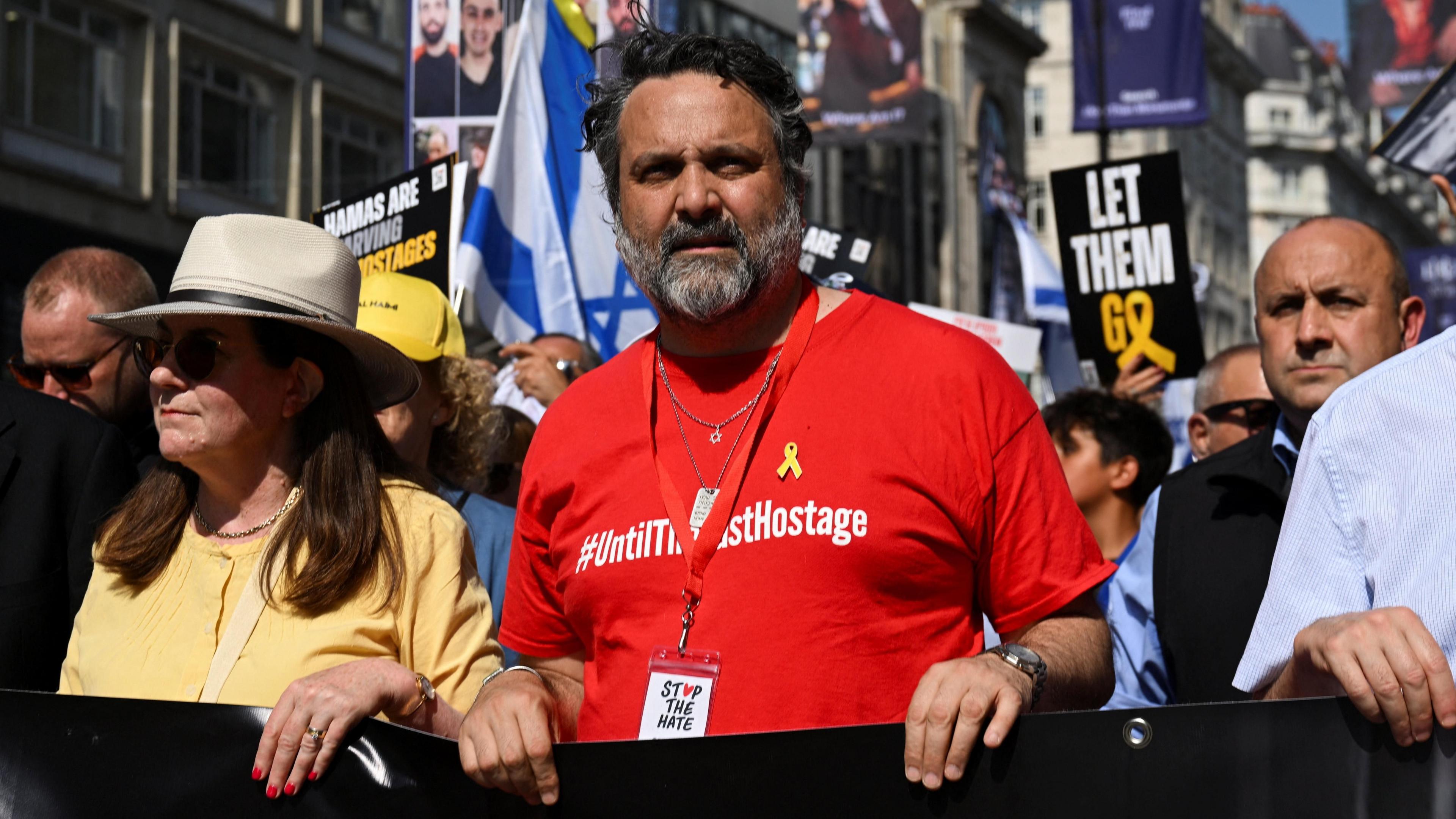 Adam Ma'anit, a man with short black hair and greying facial hair wearing a red t-shirt, walks at the front of a large protest with an Israeli flag and several placards in the background, including one which says 'let them go'.