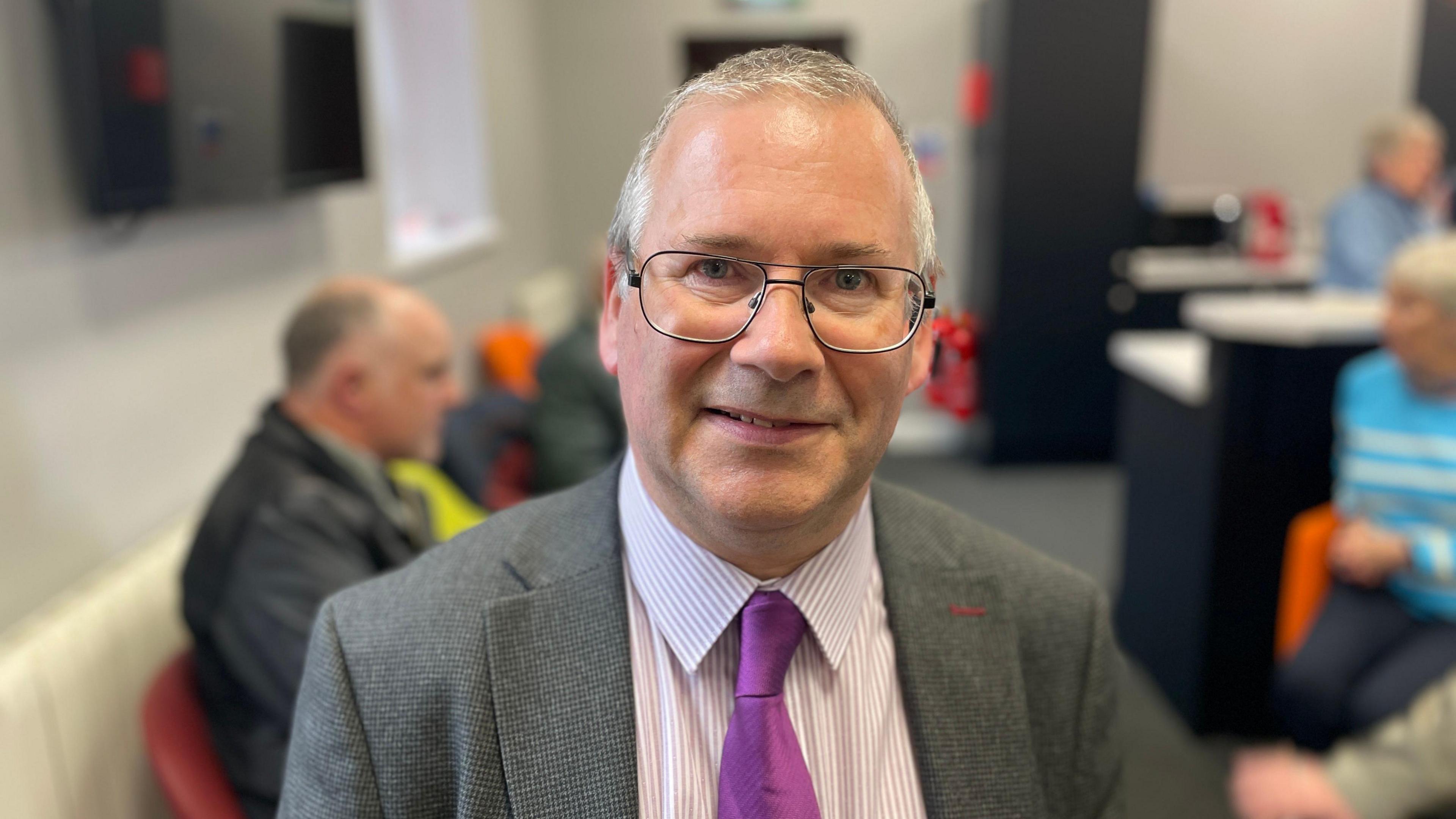 Dr David Hume looking and smiling a camera. He is wearing glasses, grey blazer, purple tie and pin-stripe shirt