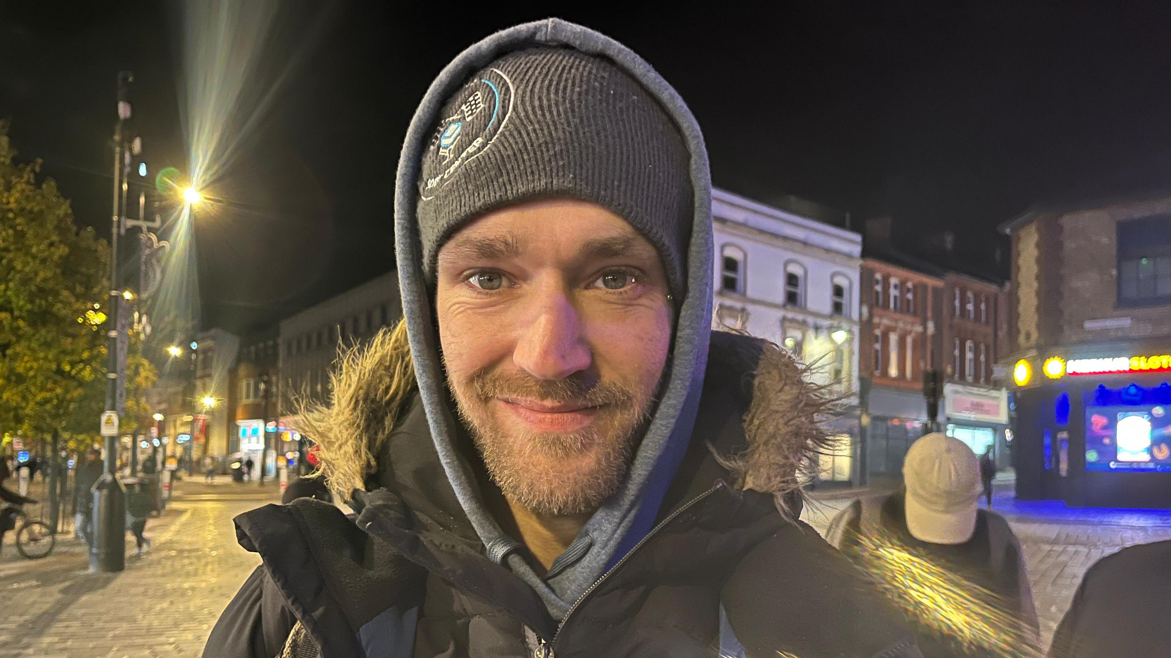 Jamie who is homeless smiles as the camera as he stands outside in Luton town centre after dark. There are buildings and street lights behind him, as well as a person with a baseball cap whose head is down. Jamie wears a grey parka coat with a furry edged hood, a beanie hat and a grey hoodie with his hood up. He is white and has grey eyes and a neat goatee beard/moustache. 