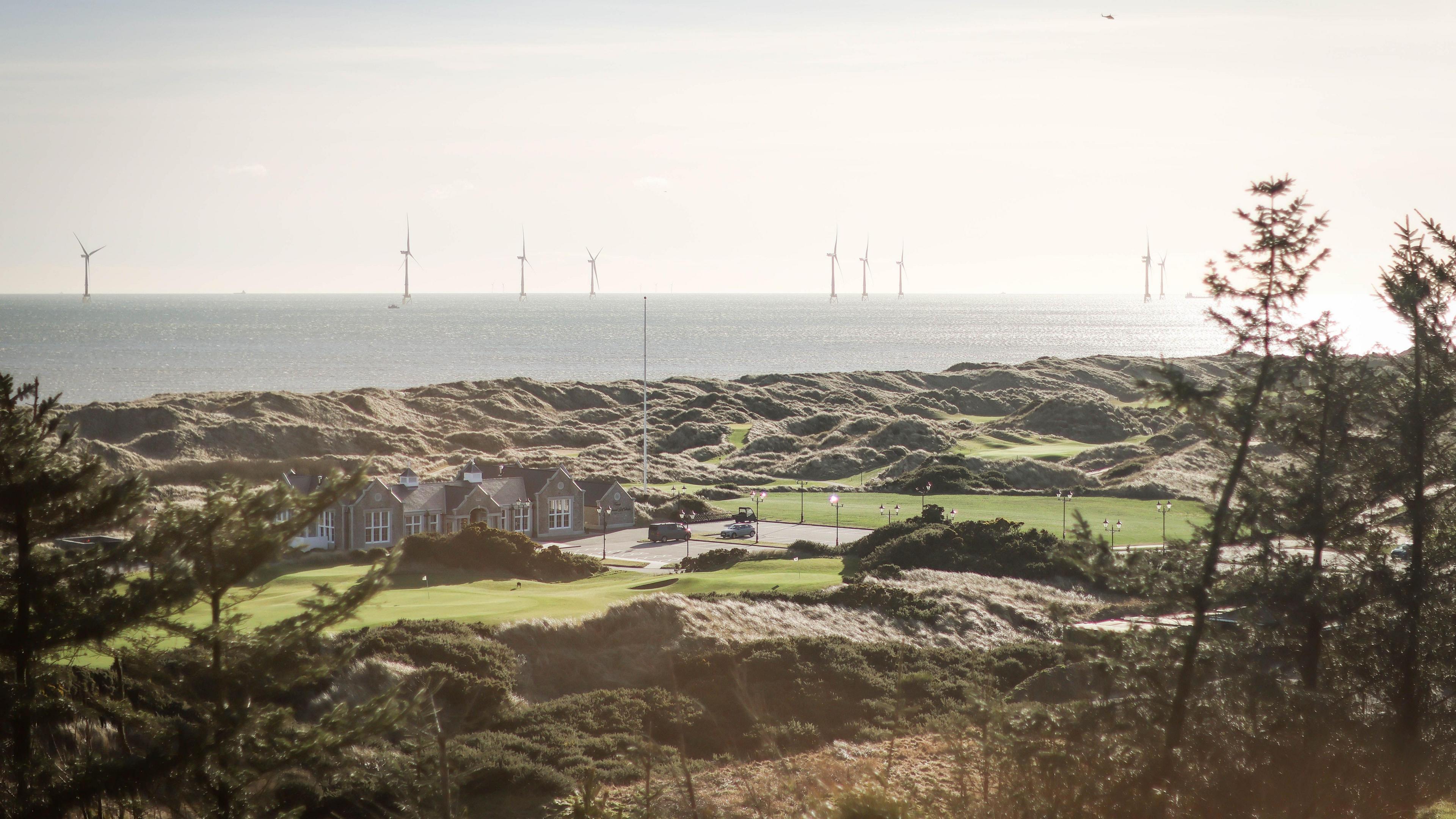 A golf course with a club house and greens next to the North Sea, which has wind turbines in the distance.