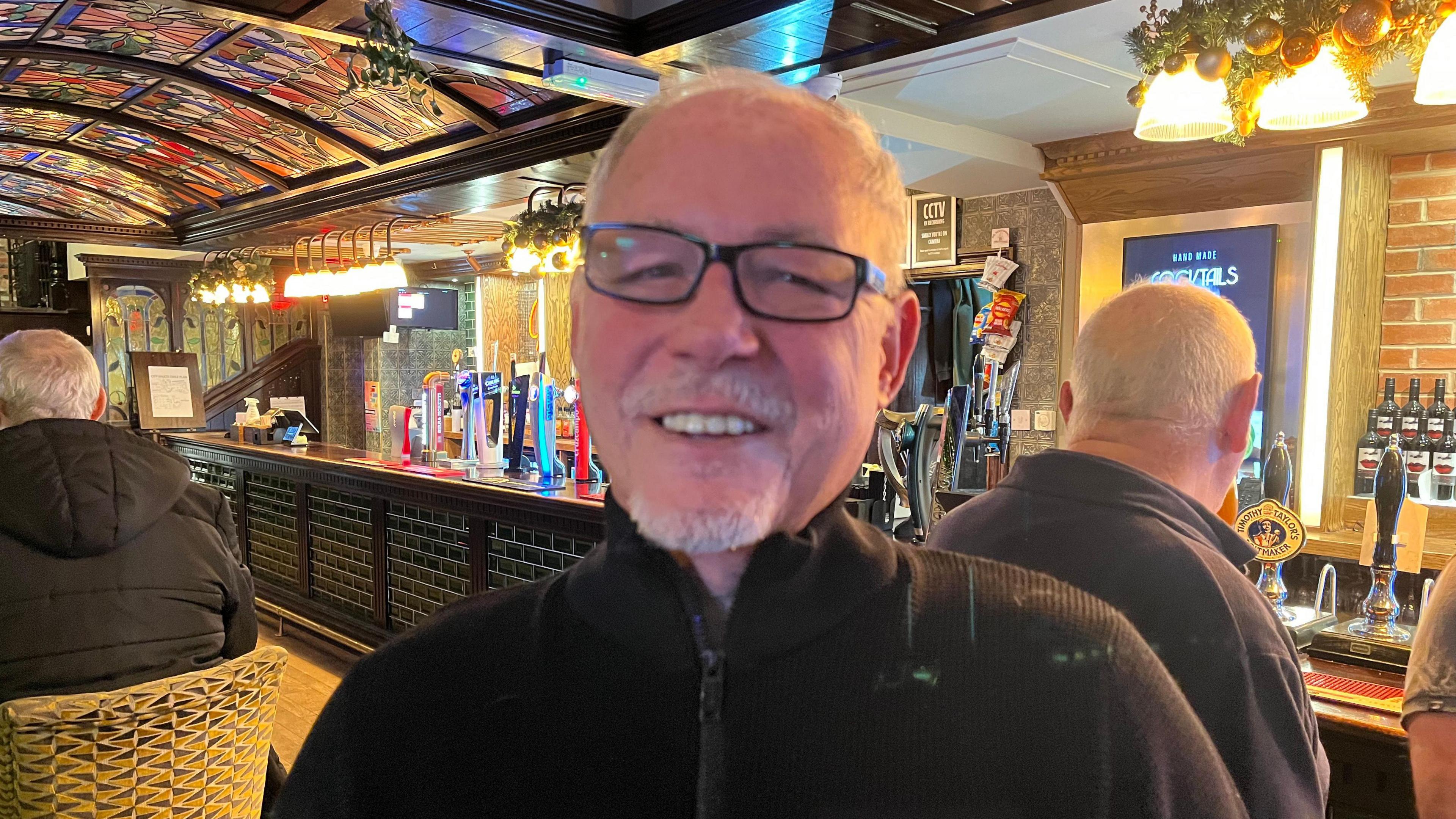 White man with thinning hair wears dark rimmed glasses and a black sweater and stands in front of a well-lit pub bar complete with beer pumps