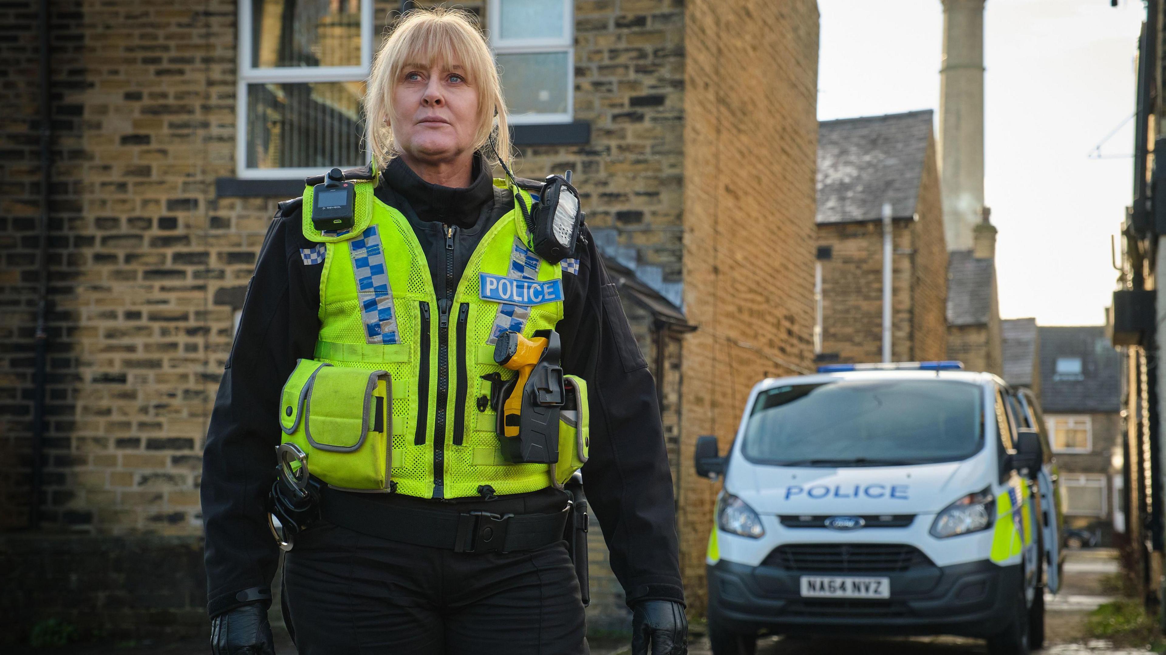 Blonde-haired Sarah Lancashire in her role as Sergeant Catherine Cawood stands in a police uniform and a hi-vis green jacket, which has a radio and a Taser attached to it, in front of a sandstone-built terraced house and a police van parked in the alley beside it