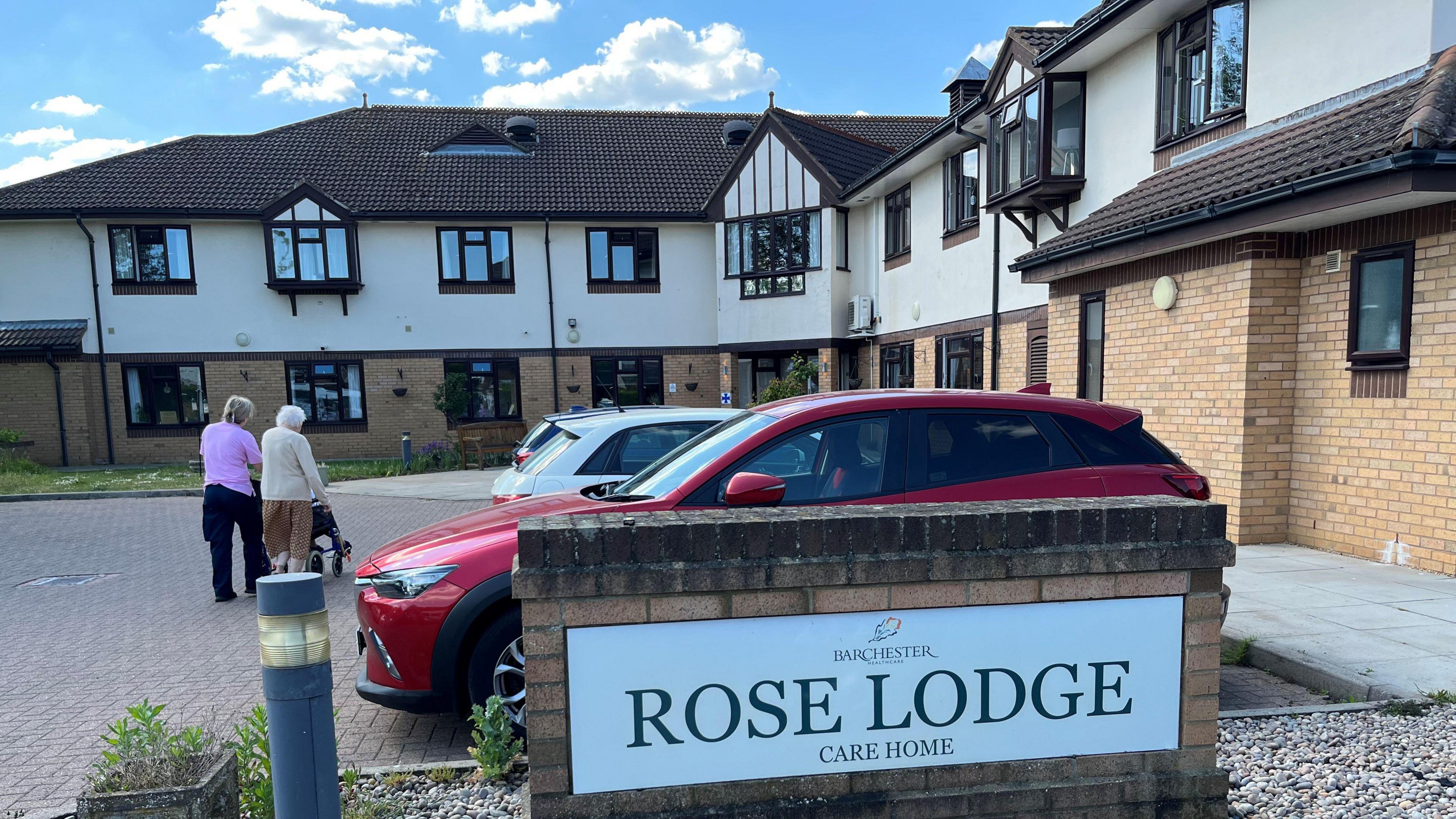 The Rose Lodge Care Home front entrance, large two-storey building, fairly modern, lower half brick, upper section render, magnolia colour, there are cars parked at the front with a couple of people walking towards the door. There is a large sign on a small brick wall saying 'Rose Lodge Care Home' on it.