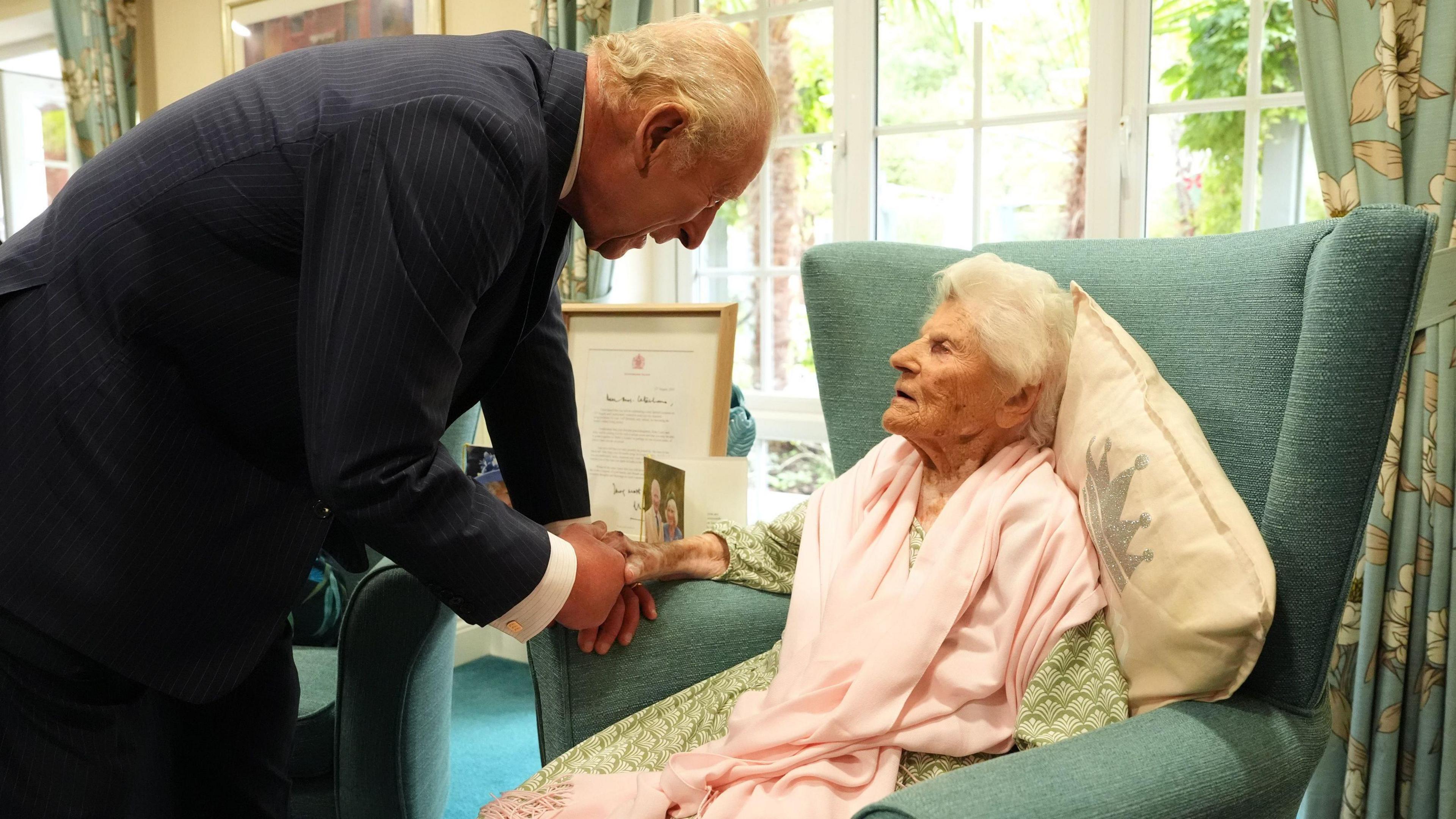 King is shown standing up and shaking hands with 116-year-old Ethel Caterham, as she sits in an arm chair with a pillow behind her. 