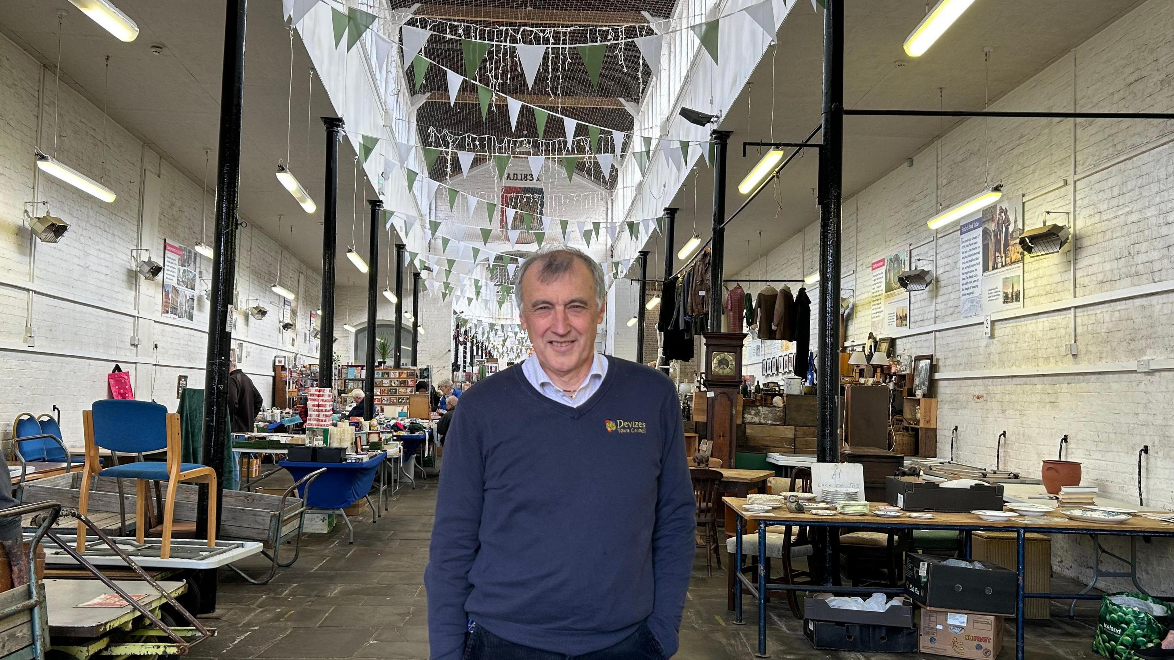 Simon Fisher smiles at the camera wearing a dark blue jumper with a town council logo. He is standing inside the market, with table-top stalls behind him and bunting between the columns.