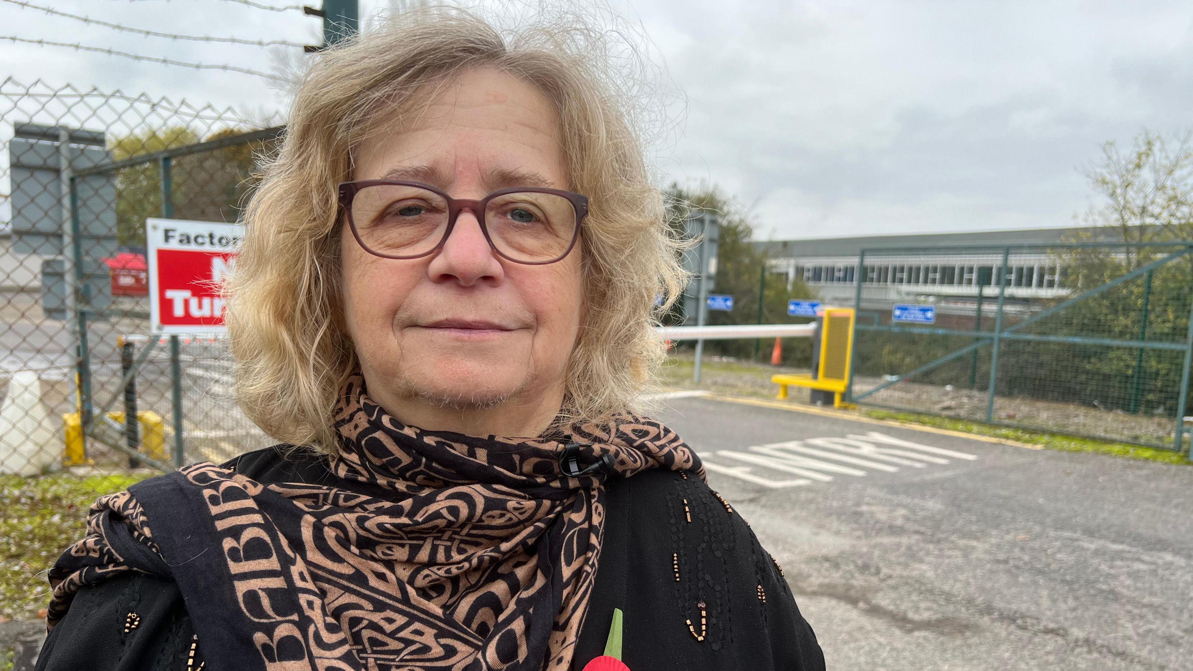 Councillor Chris Willmore has short blonde hair and dark brown glasses. She wears a black top, a black scarf with a brown print and a poppy on her chest. She stands outside the Hotpoint factory.