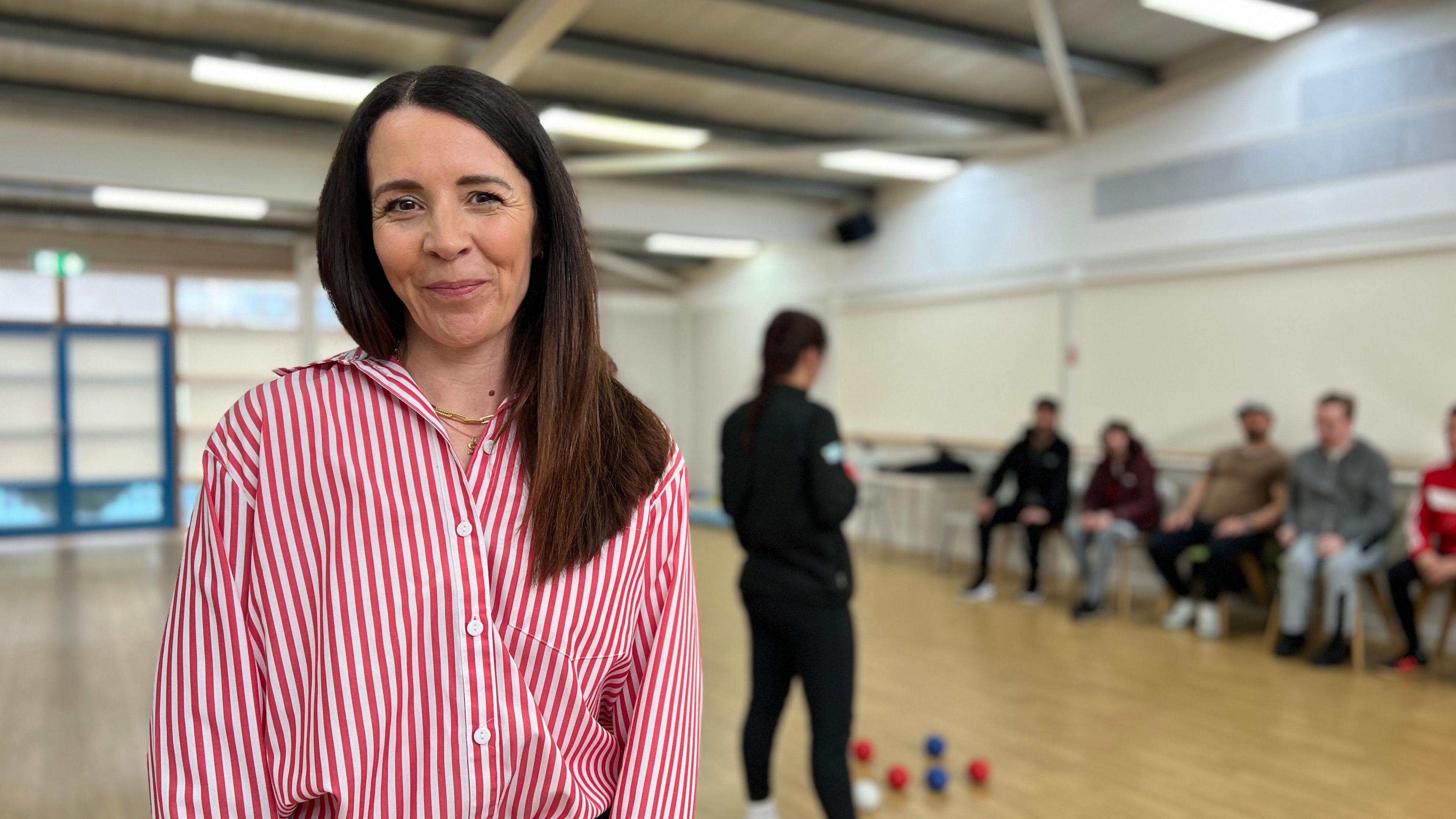 Sarah McCluskey smiles in front of the camera with people playing indoor bowls behind her