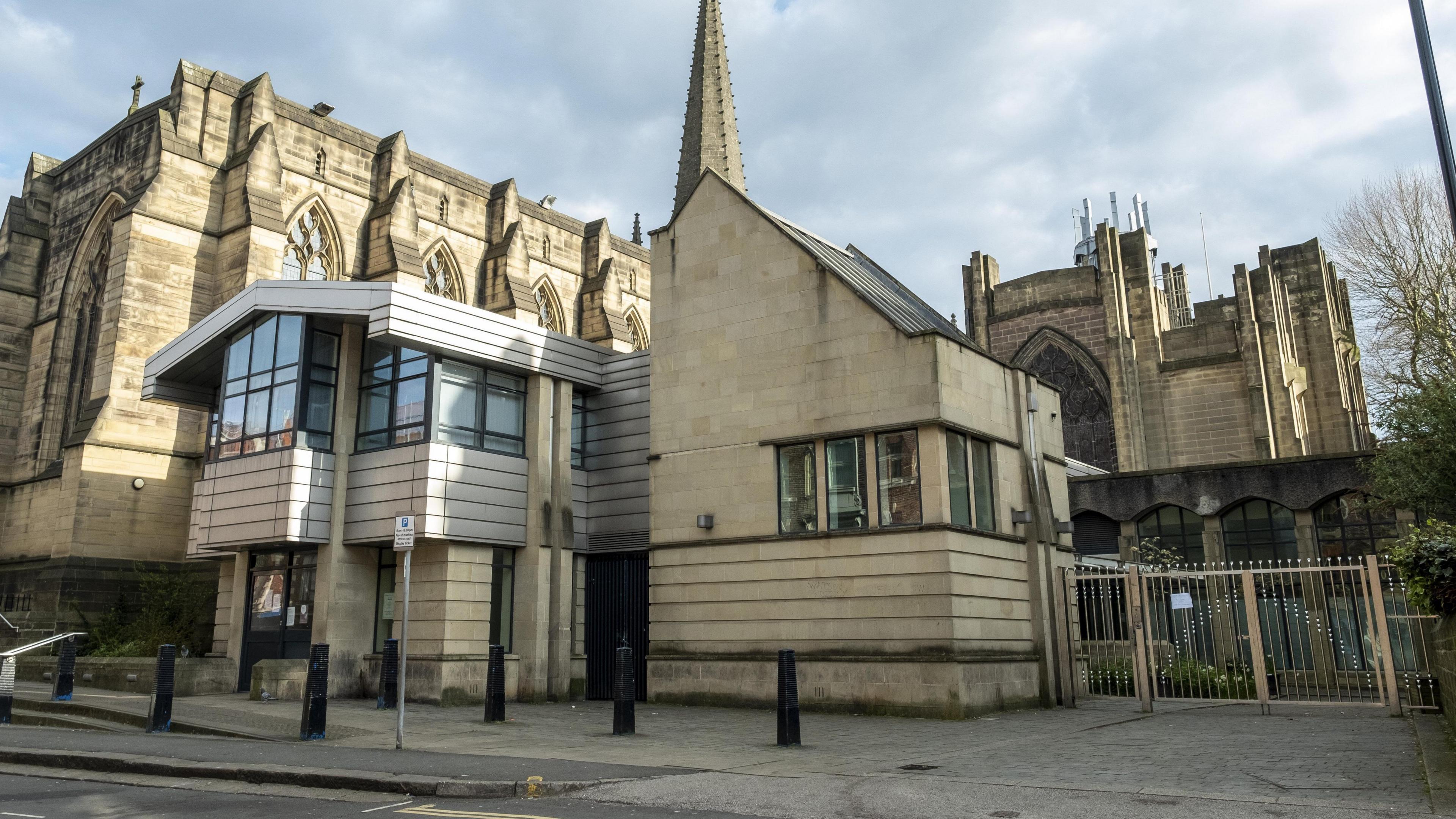 A stone building with metal gates stands infront of a cathedral.