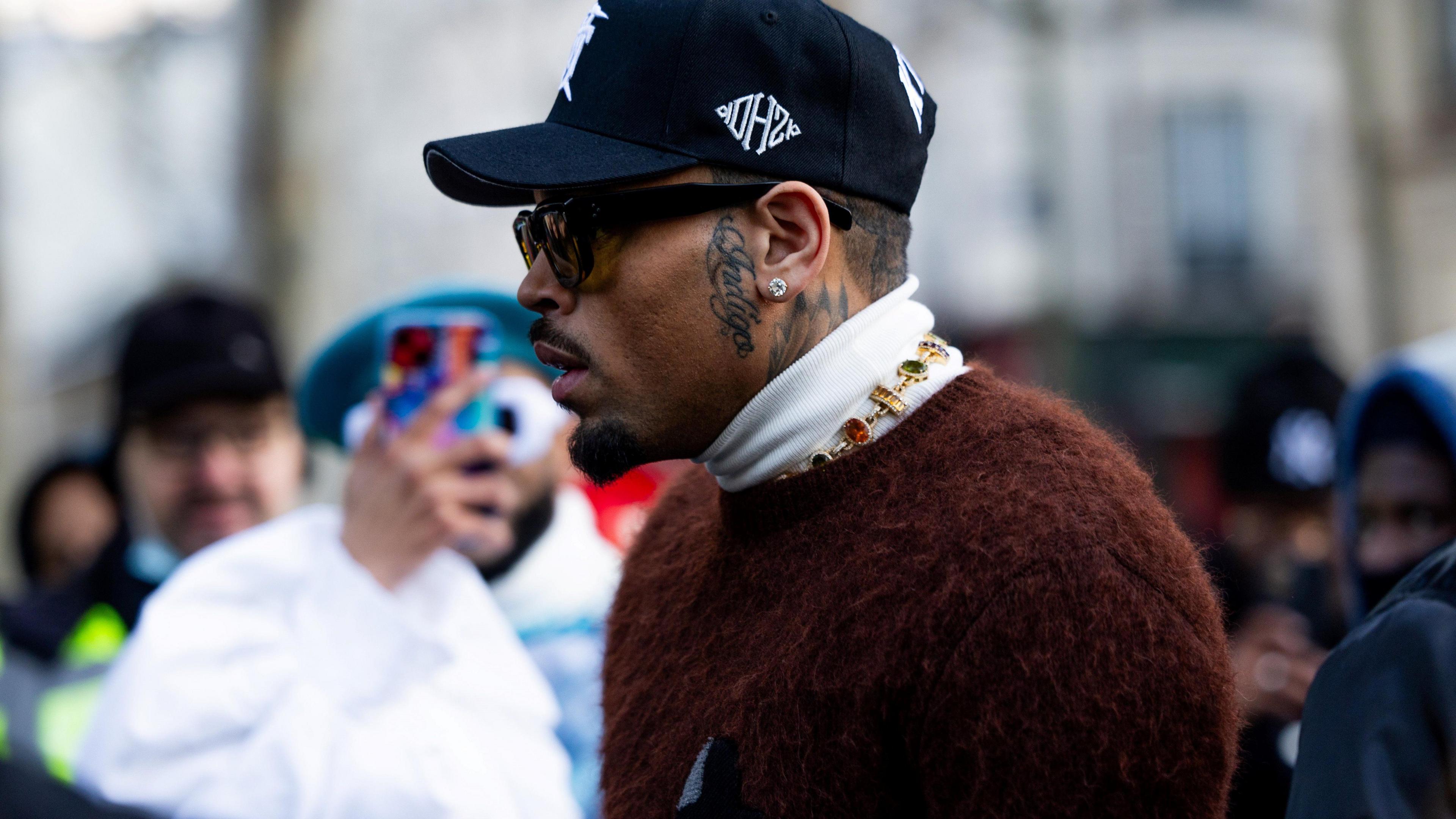 Chris Brown at Paris Fashion Week in January 2024. He wears a black baseball cap and dark sunglasses with a fluffy brown cardigan over a white turtle-neck jumper. Fans can be seen trying to take photos of him in the background. 