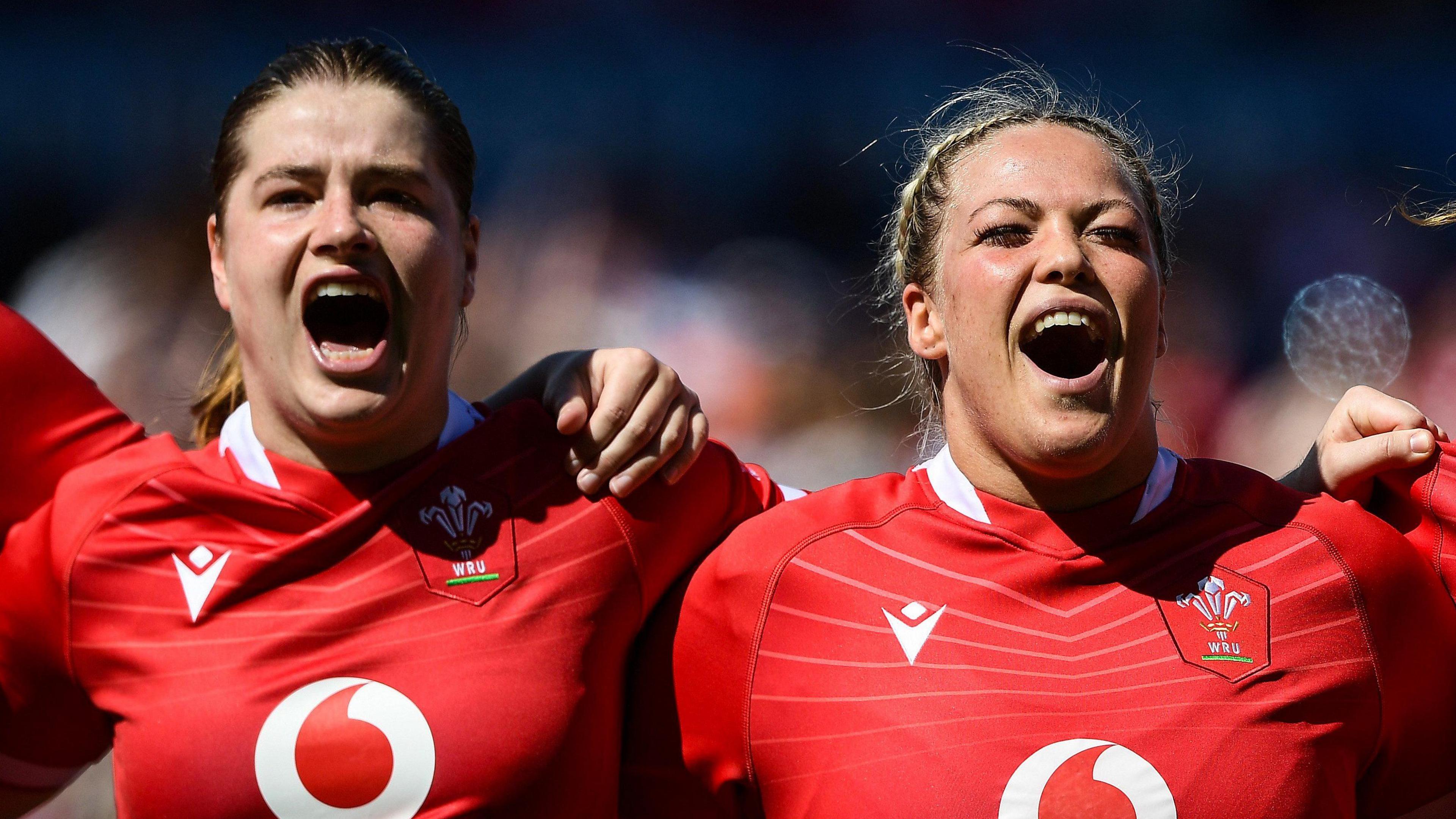 Bethan Lewis and Kelsey Jones wearing Wales kit standing in a line with their arms around each other during the national anthem of a Wales match