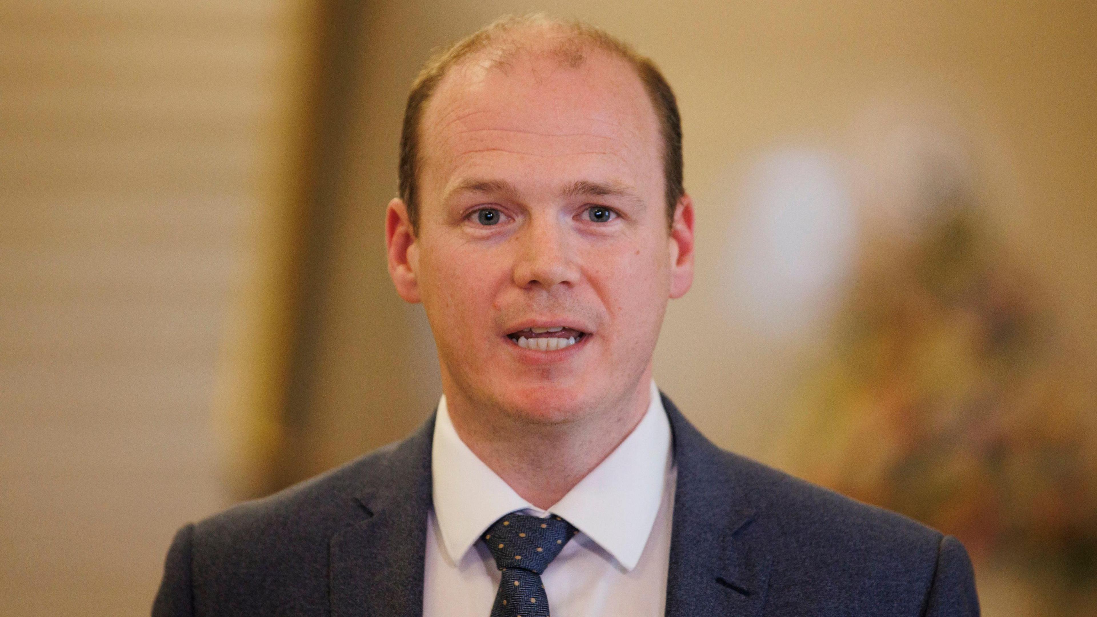 A man with balding dark hair, a white shirt dotty tie and navy suit speaks