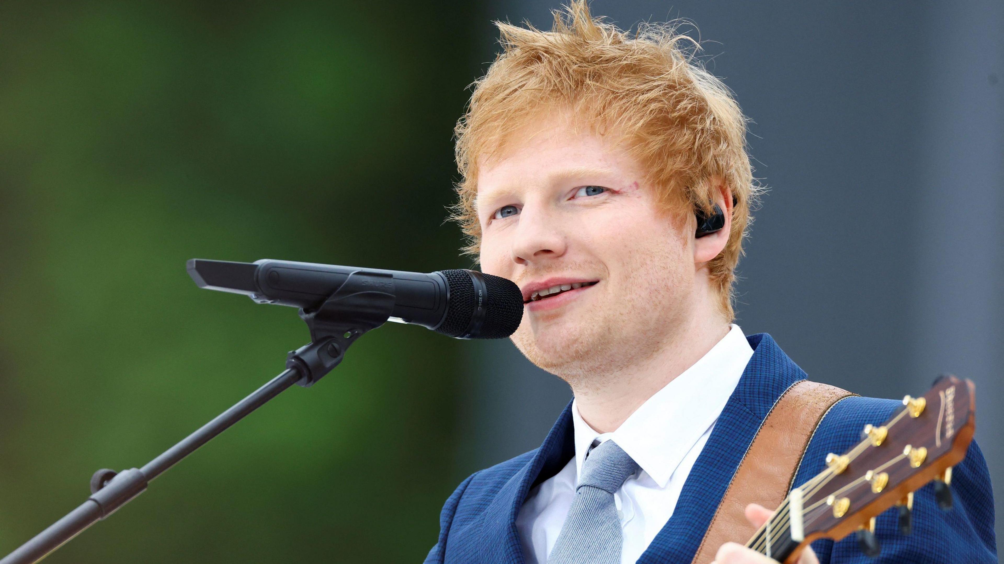 Ed Sheeran a man who is standing outside, wearing a navy suit with white shirt and grey tie. He is singing into a microphone and holding a wooden guitar.