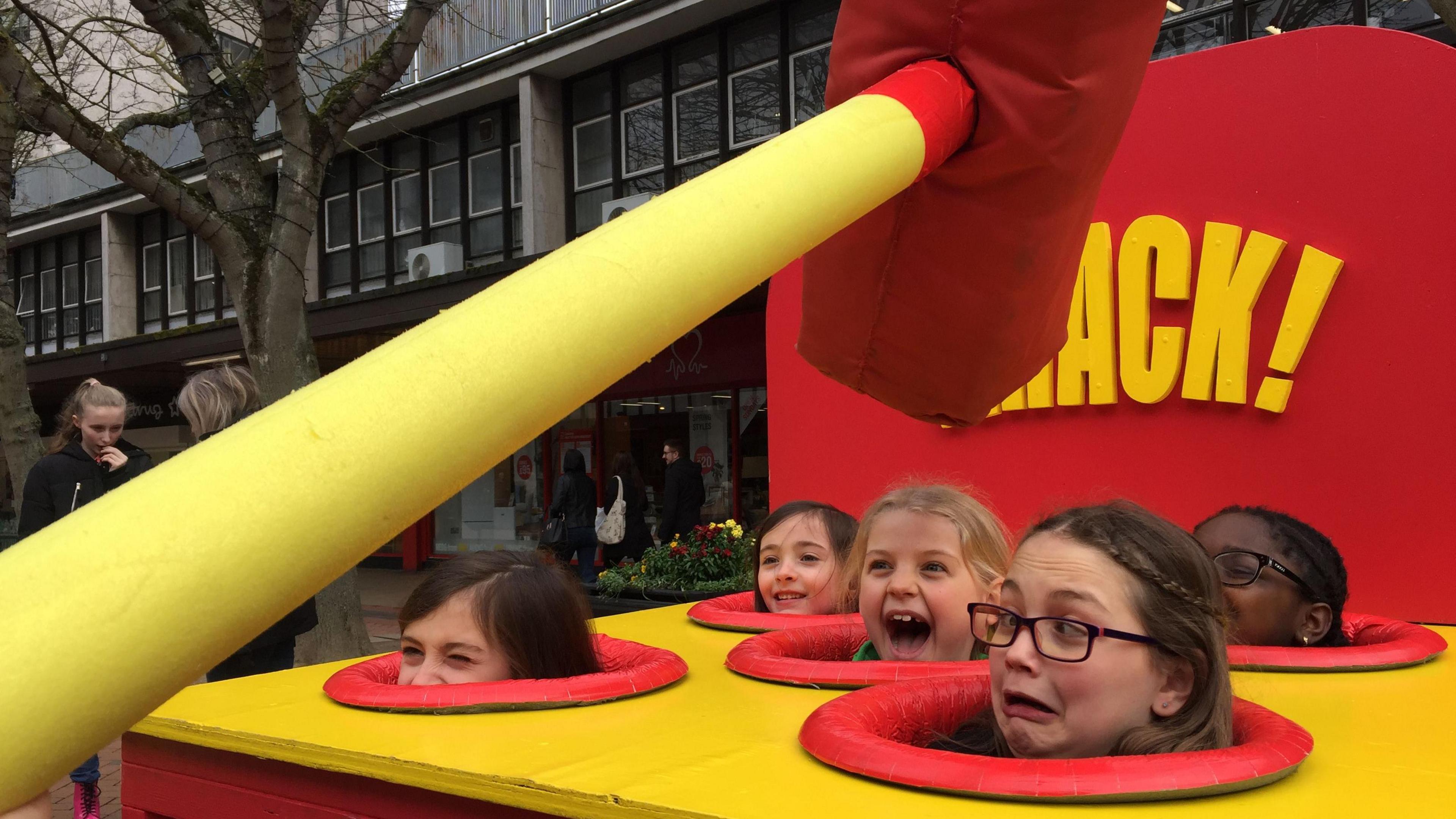 A yellow whack-a-mole stand set up with a large foam hammer raised about to descent with five scared children looking on