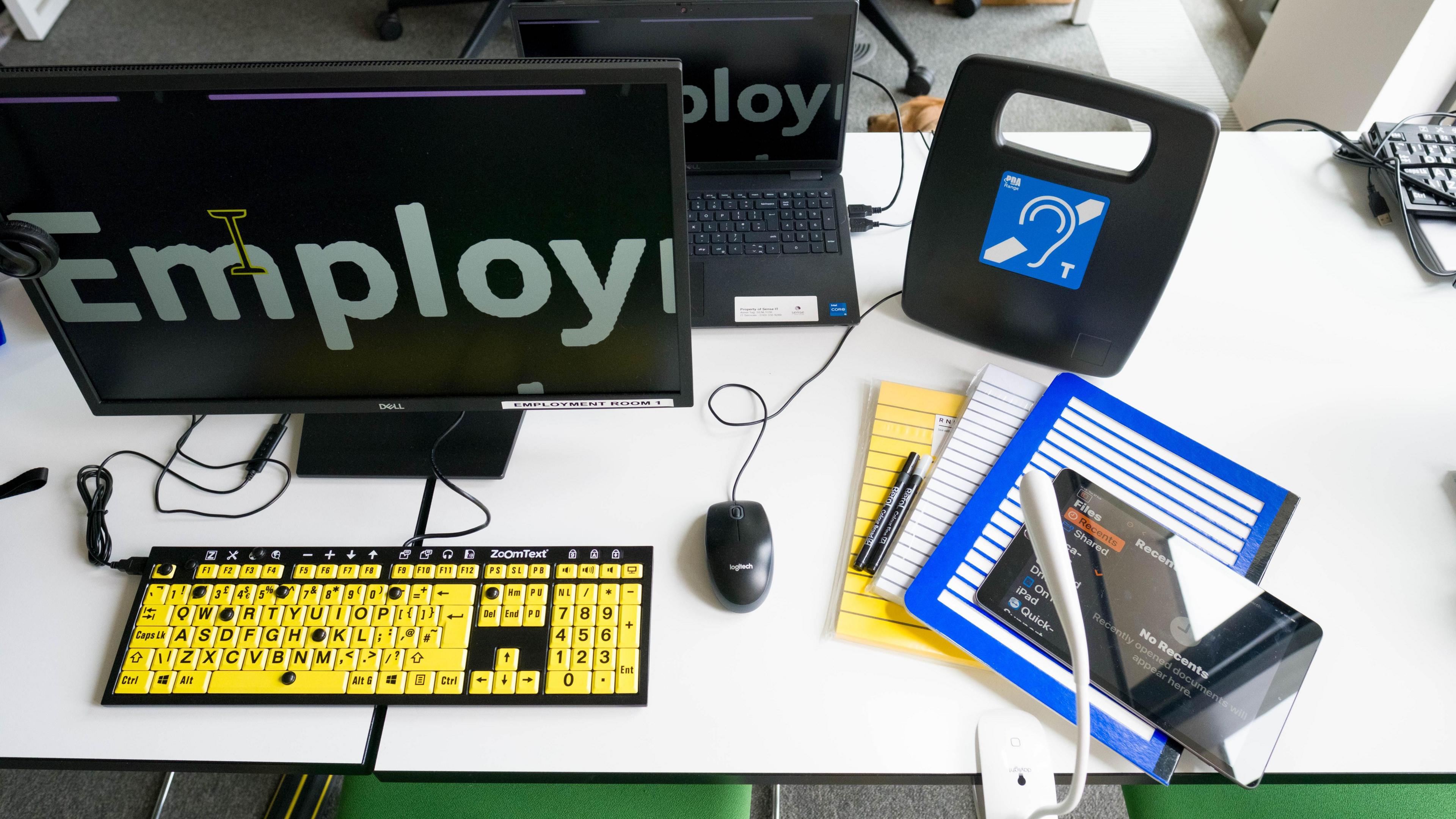 A yellow keyboard, monitor, mouse, note pads, hearing device, aids to help people in the work place, on a desk, in an office. The desk is white, with cables coming from the equipment. 