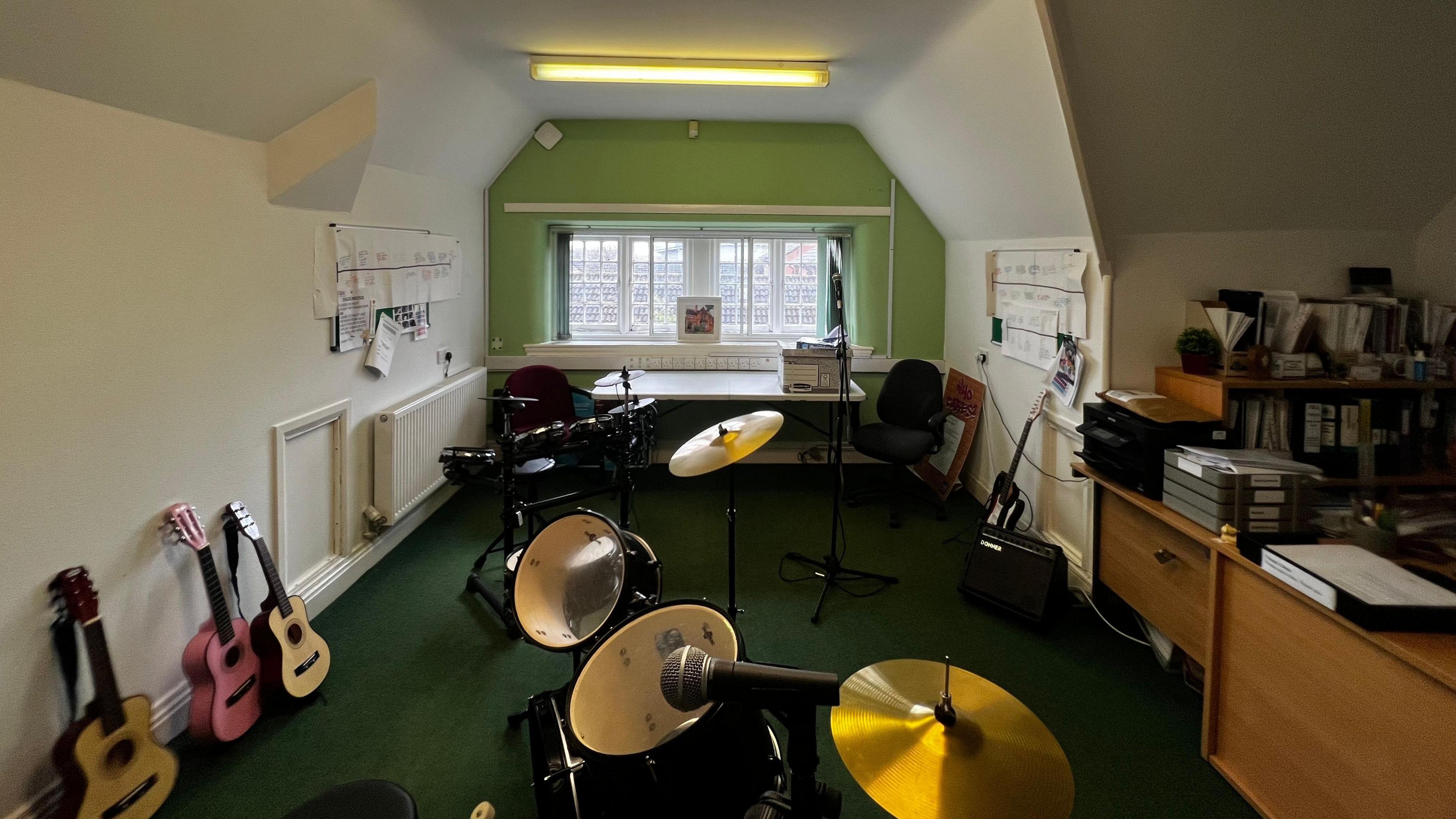 A room that is cluttered with instruments filling up most of the floor space, with a desk off to the right hand side. The room has dark green carpet and a lime green wall at the end, surrounded by white walls