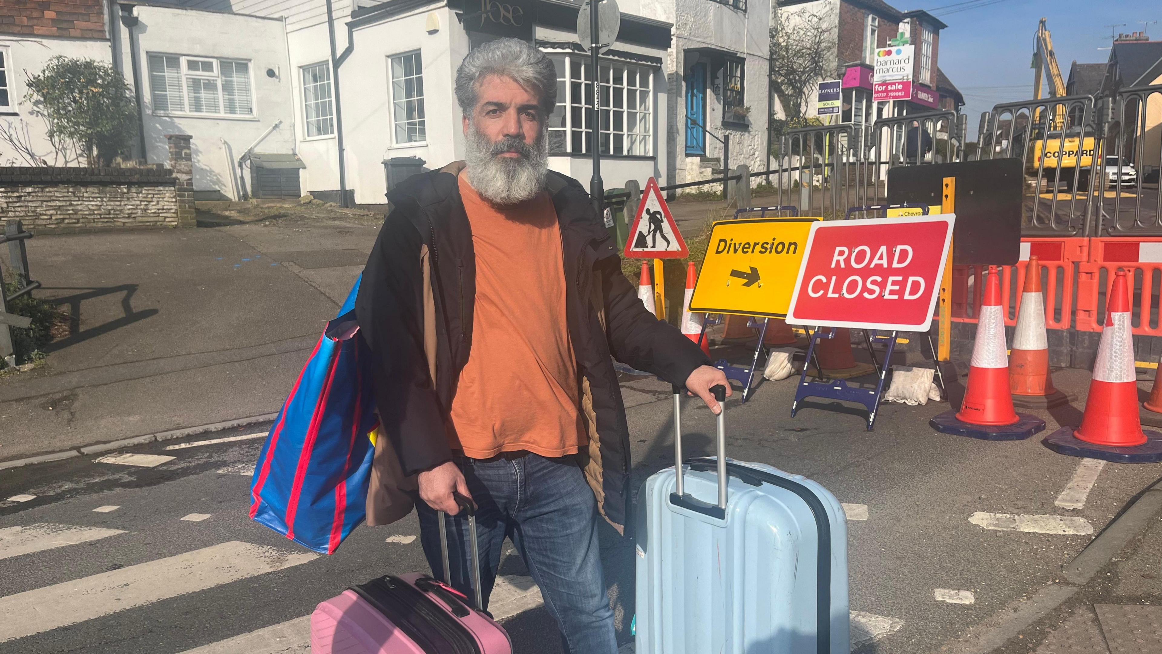 Rez Mira wheels two suitcases and carries a bag over his shoulder. He wears a brown cardigan, orange t-shirt and blue jeans, and has grey hair and grey beard. Behind him are signs saying road closed and diversion.