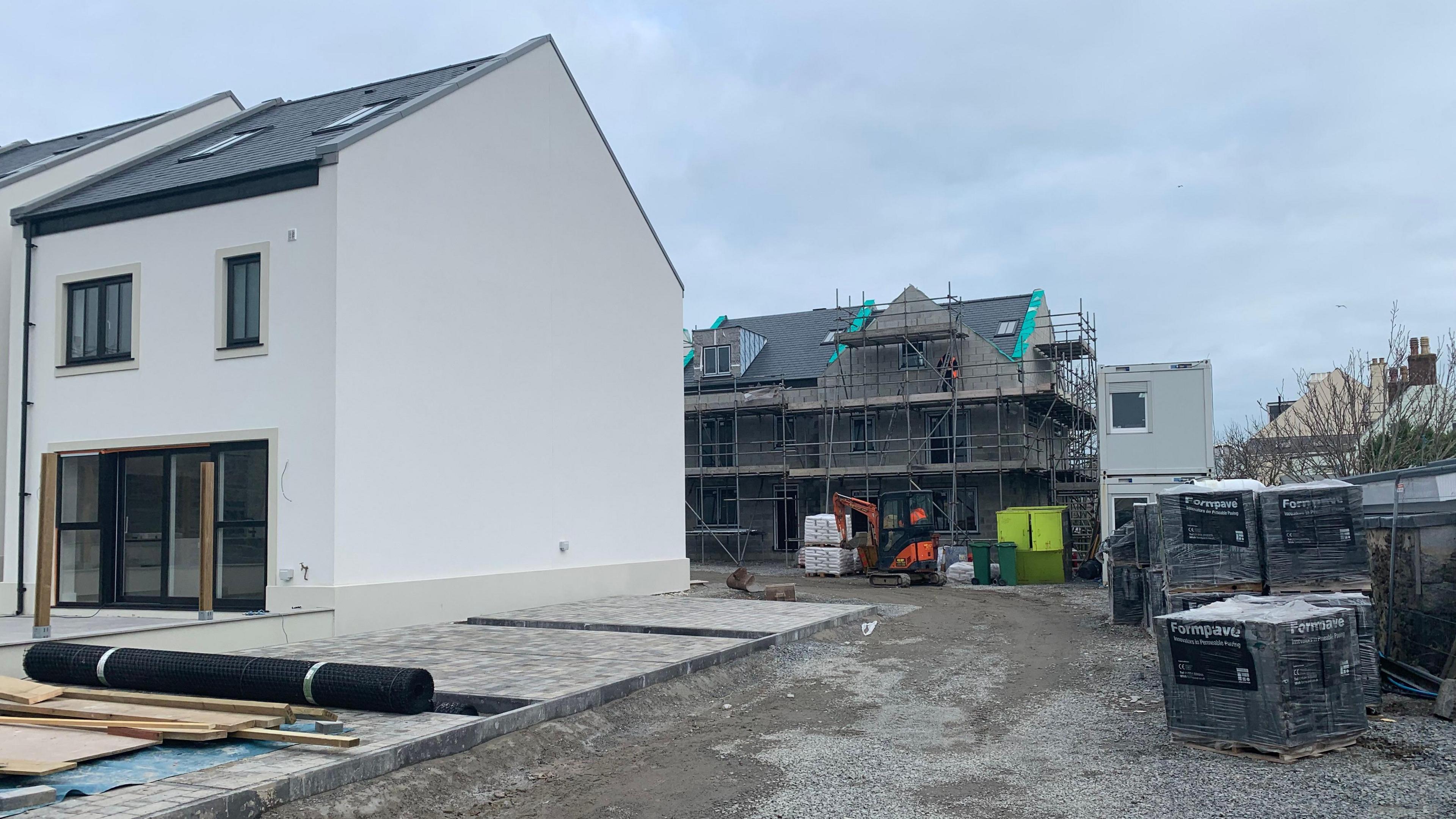 A grey house with a blue-grey roof next to a house under construction surrounded by scaffolding next to piles of grey blocks. 