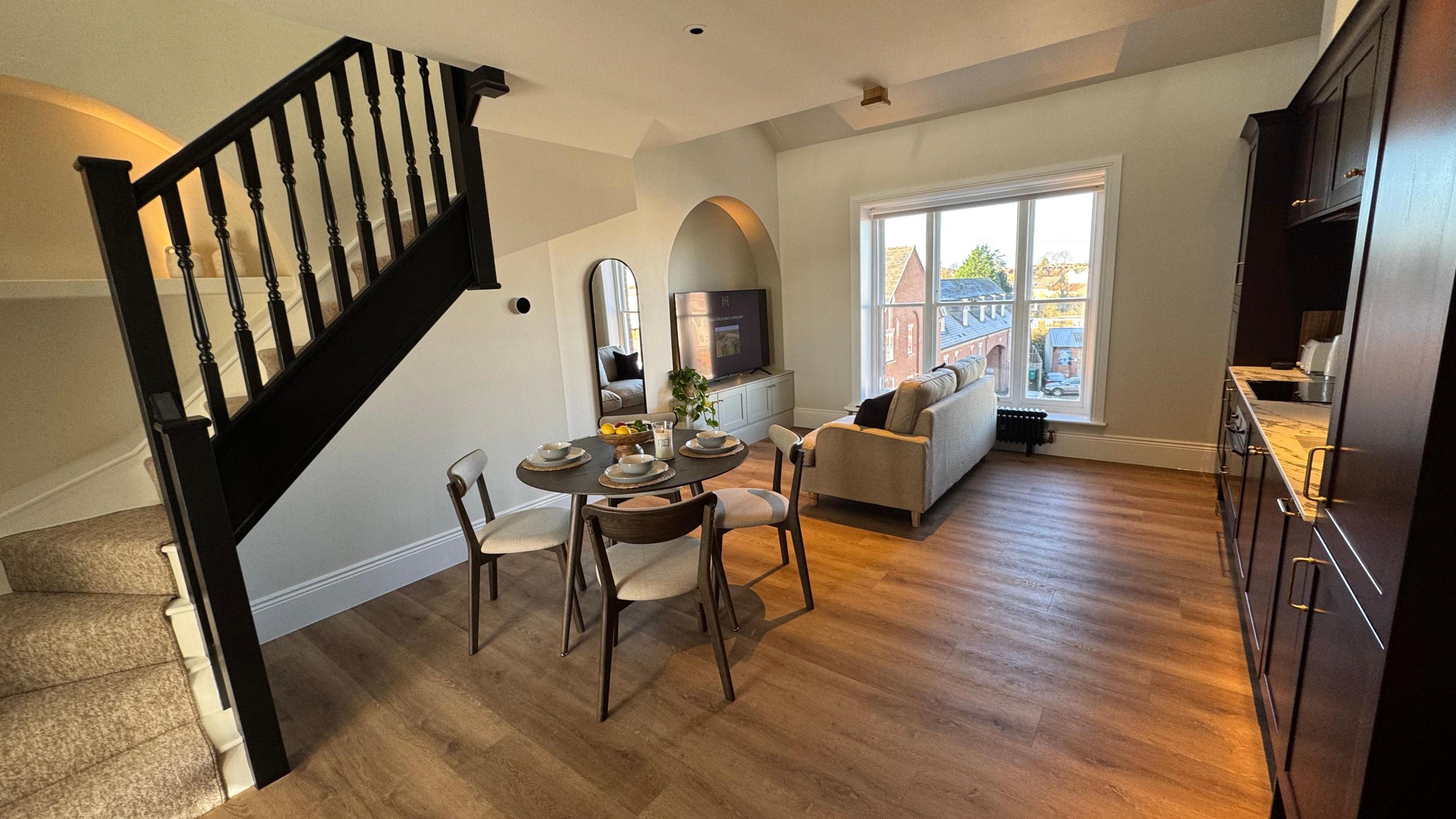 A room with wooden flooring, a staircase with brown wood banister. There is a small table with three chairs around it, a sofa, television and window looking out onto building in the street below