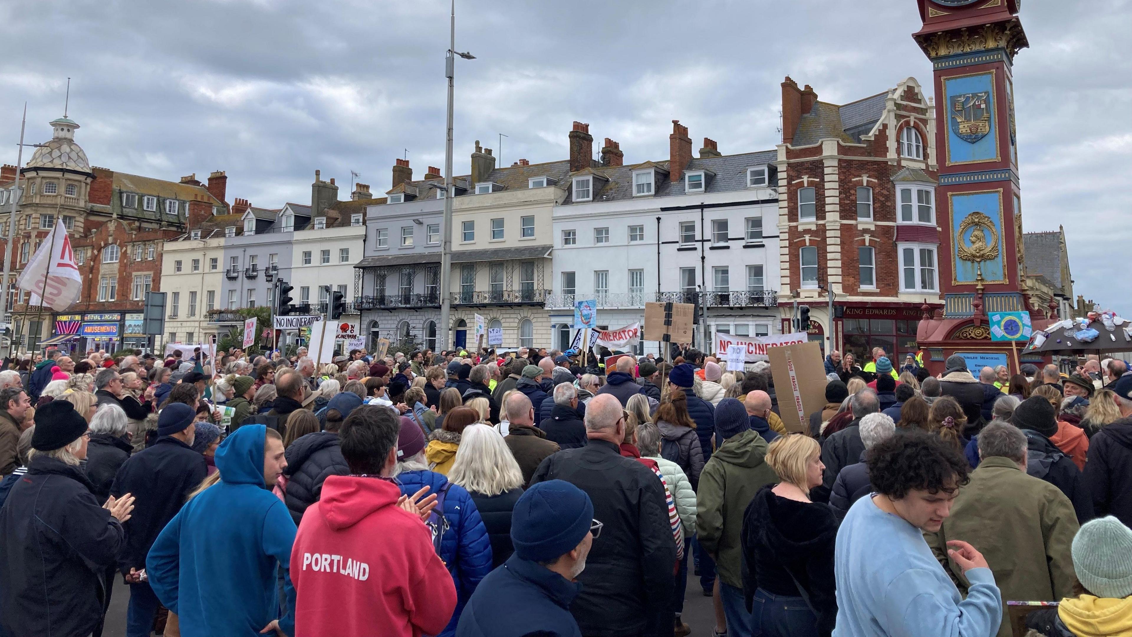 A crowd of several hundred people standing in a large open part of a town, with a row of buildings and a clock tower behind them.