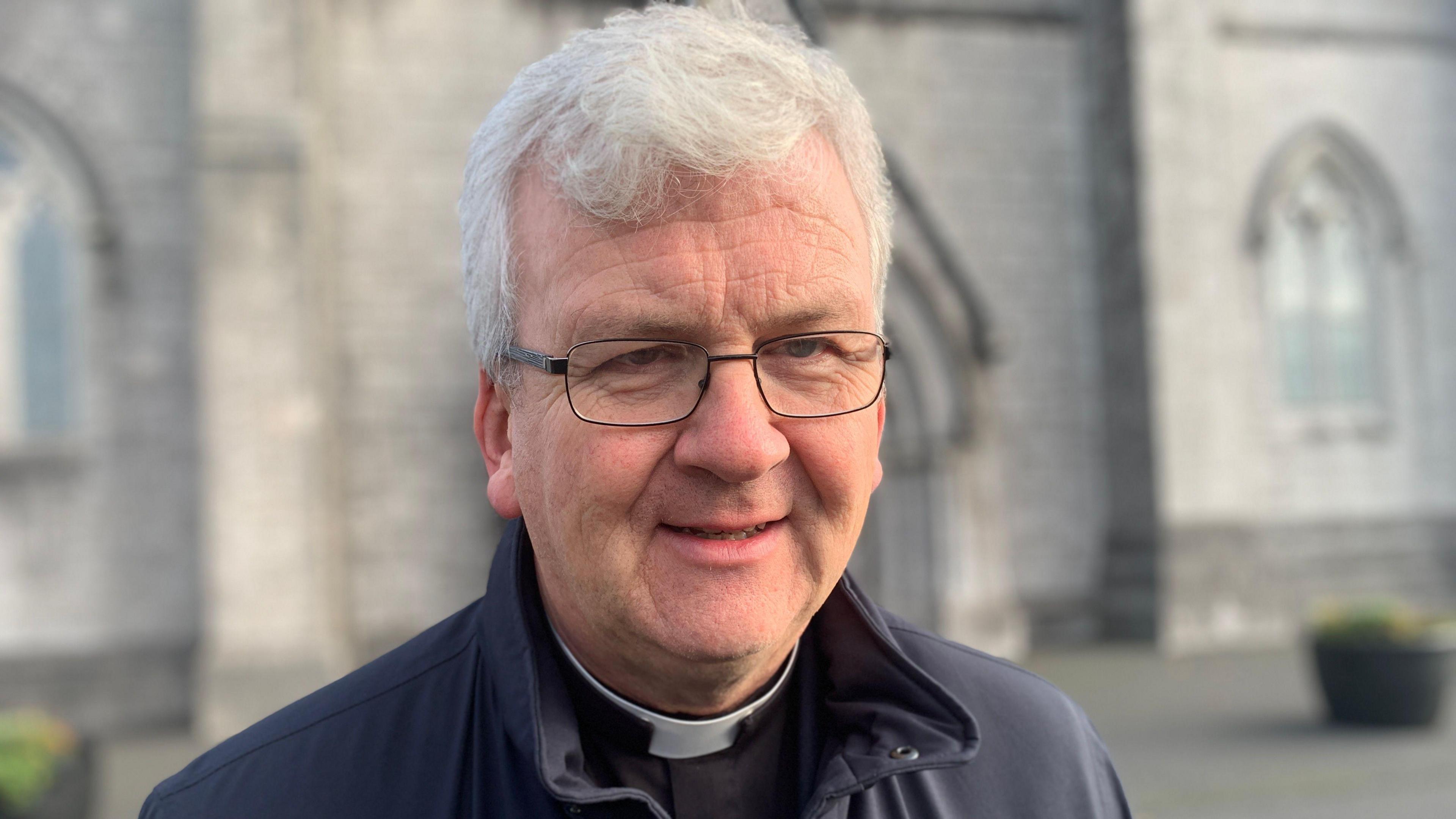 Monsignor Shane McCaughey. He has white hair, wearing glasses, a clerical collar and black zip up rain jacket. The chapel is blurred in the background.