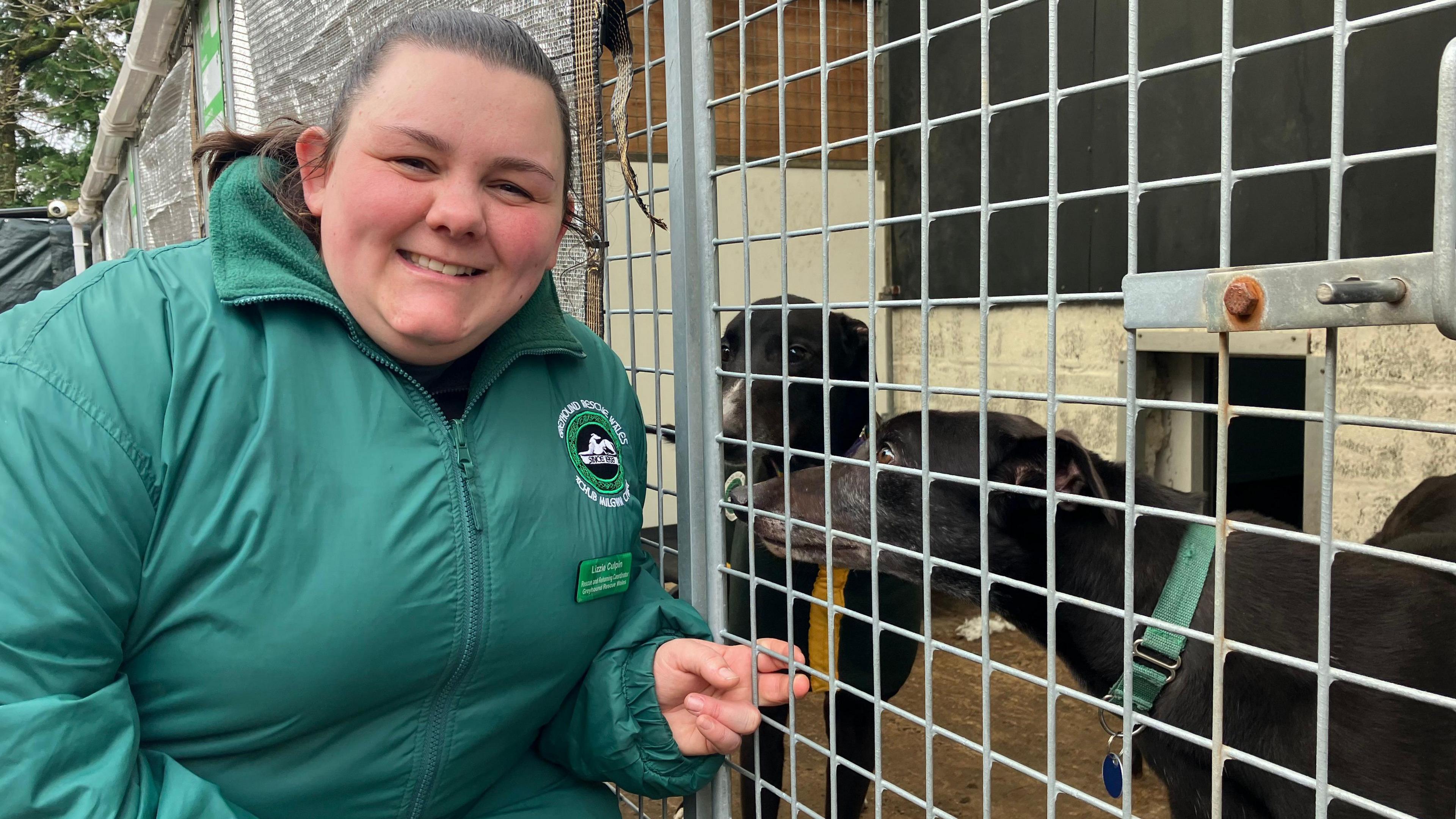 Lizzie Culpin smiles with a pair of greyhounds next to her in a cage. She is wearing a green jacket, with her dark hair tied back. 