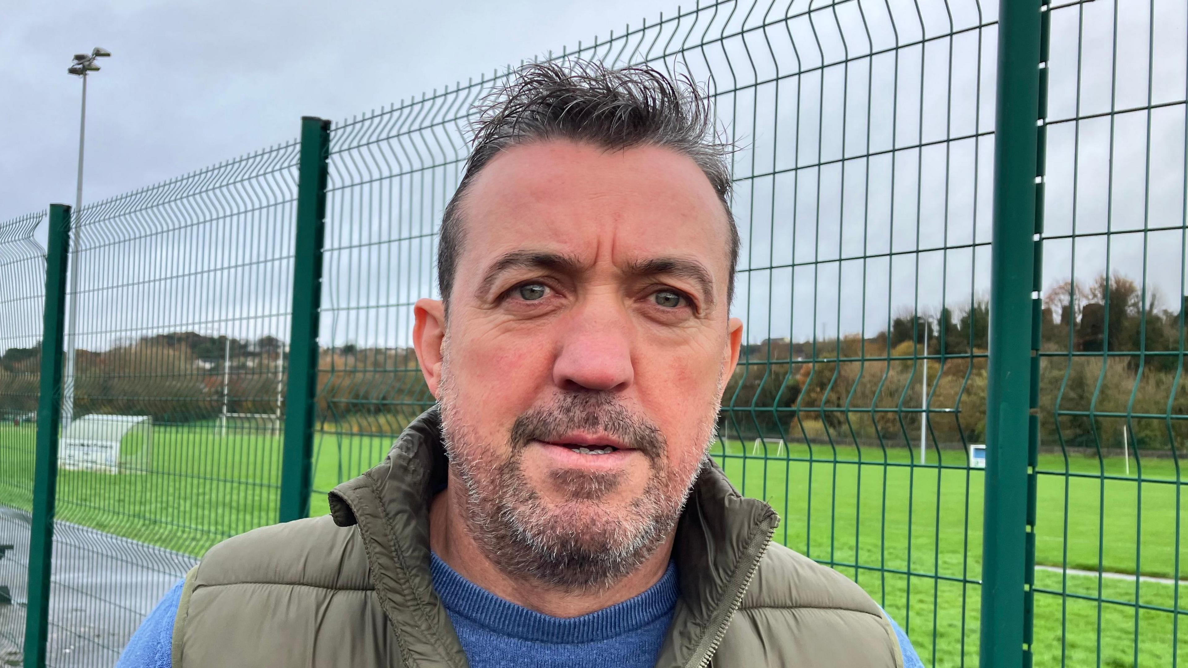 Shows a man with a beard standing beside the fence of a Gaelic football pitch