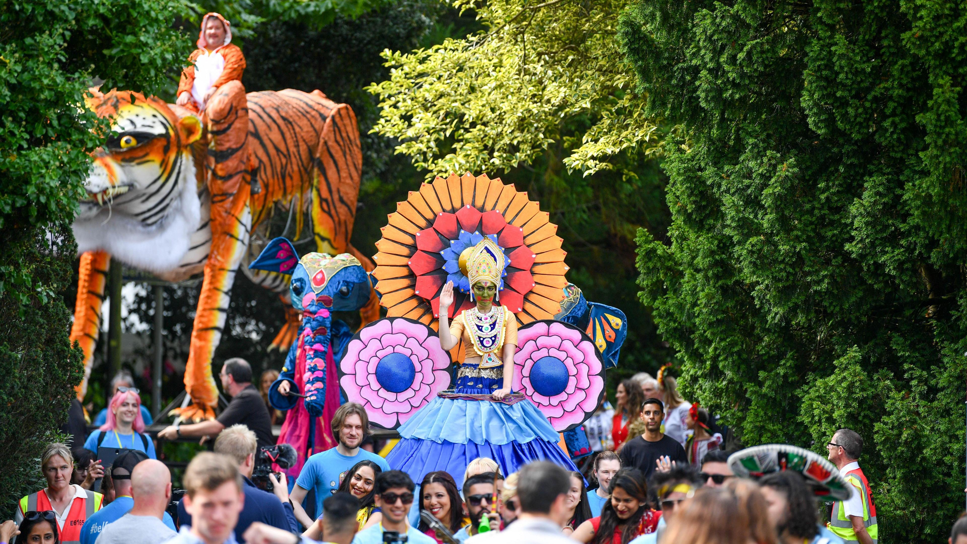 A parade is making its way through a public garden. Someone is sitting on top of a large tiger float. A crowd of people are watching. Trees are on both sides of the performers and spectators.