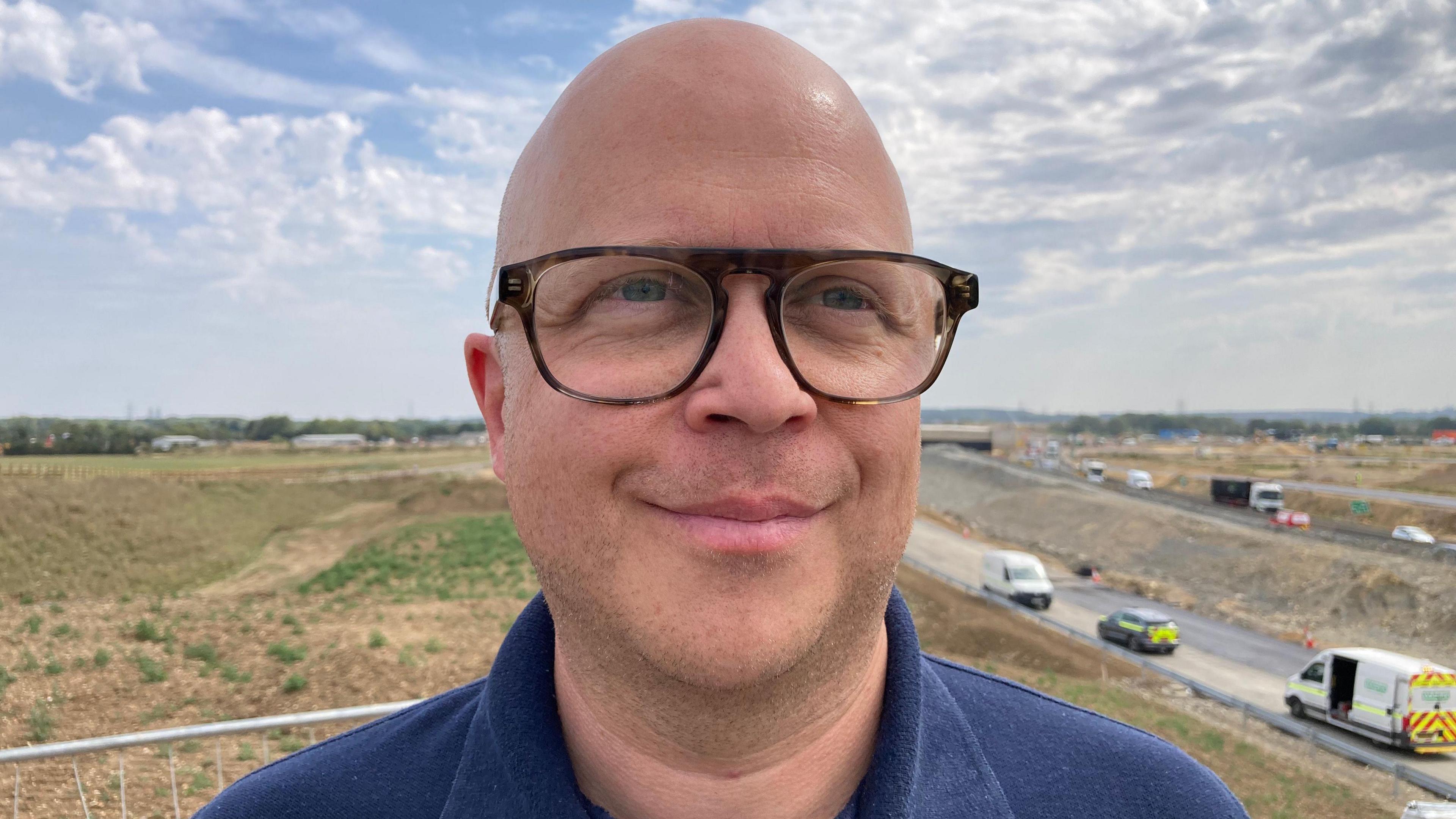 A man with glasses and a blue top is stood on a sunny day. Behind him are fields and construction workers on a nearby unfinished road.