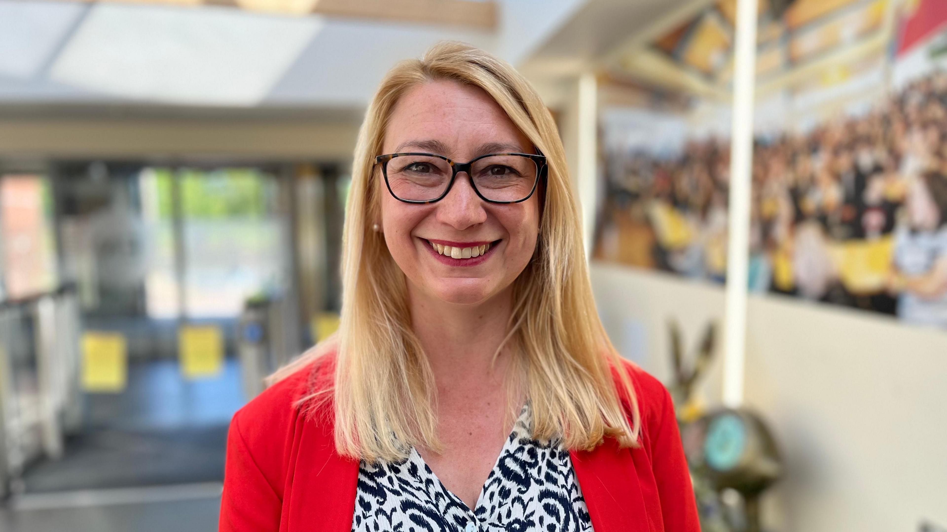 Jess Asato smiles into the camera. She has long, straight blonde hair and is wearing black glasses. She is wearing a black and white printed blouse under a red blazer.
