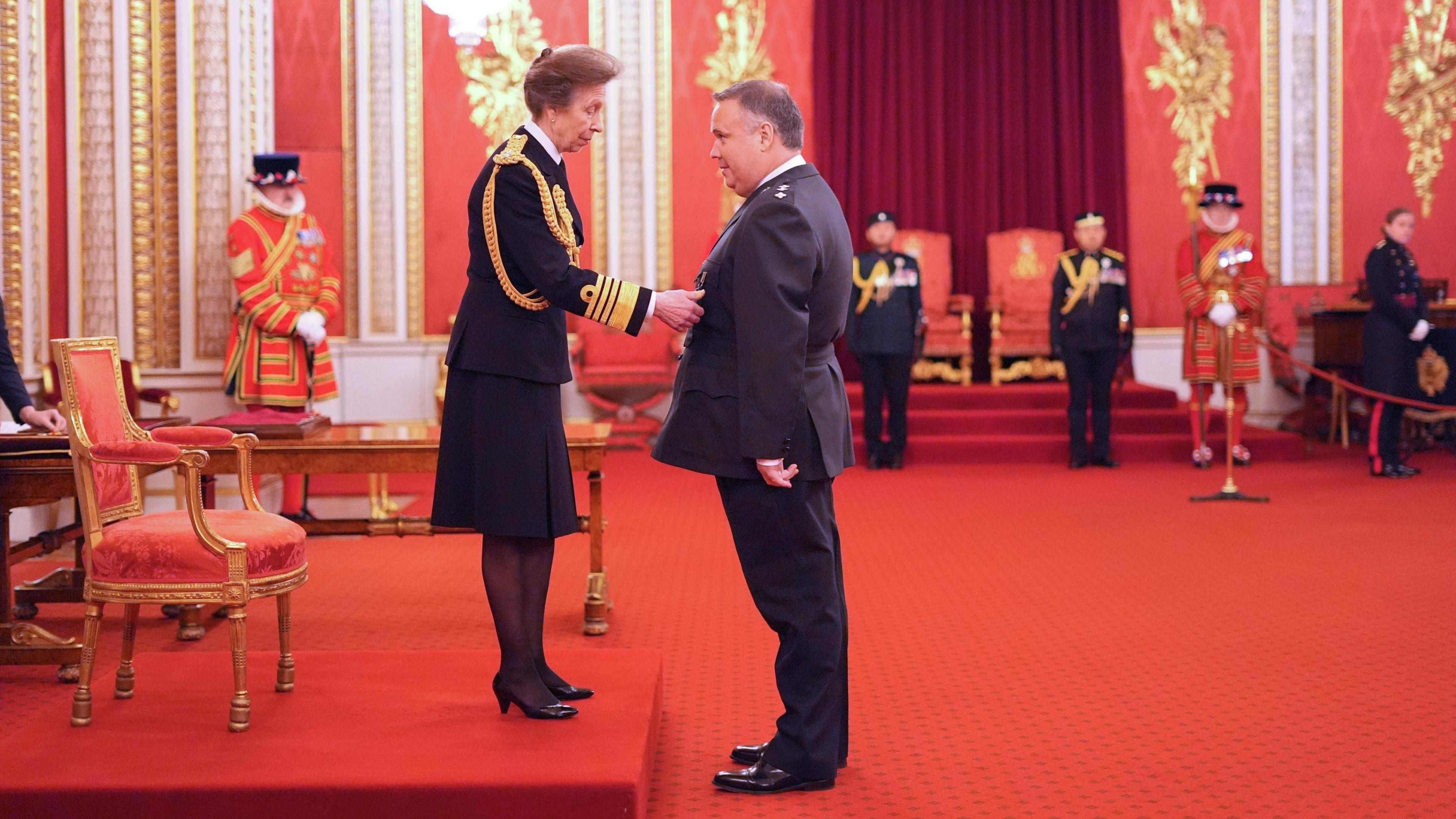 A wide shot of Princess Anne, dressed in a royal uniform, is stood on a red platform in a hall in Buckingham Palace. She is placing a medal on John Caldwell, who is stood on the right wearing a police uniform.