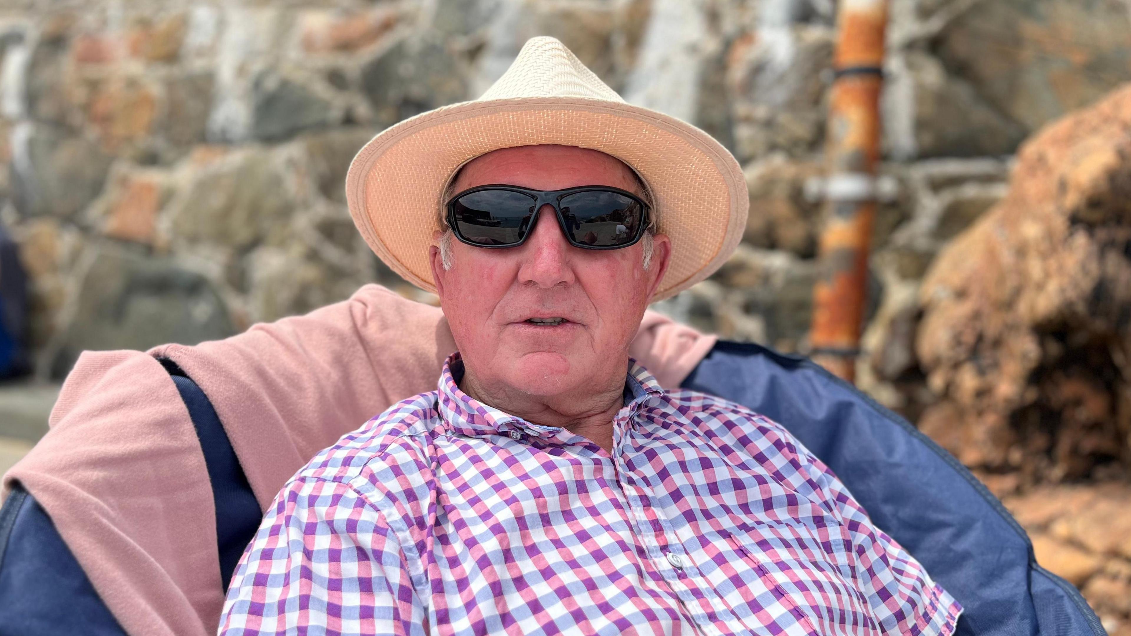 A man wearing a straw hat, black sunglasses and a red, blue and white checked top sitting down in a low chair at Guernsey's Bathing Pools. 