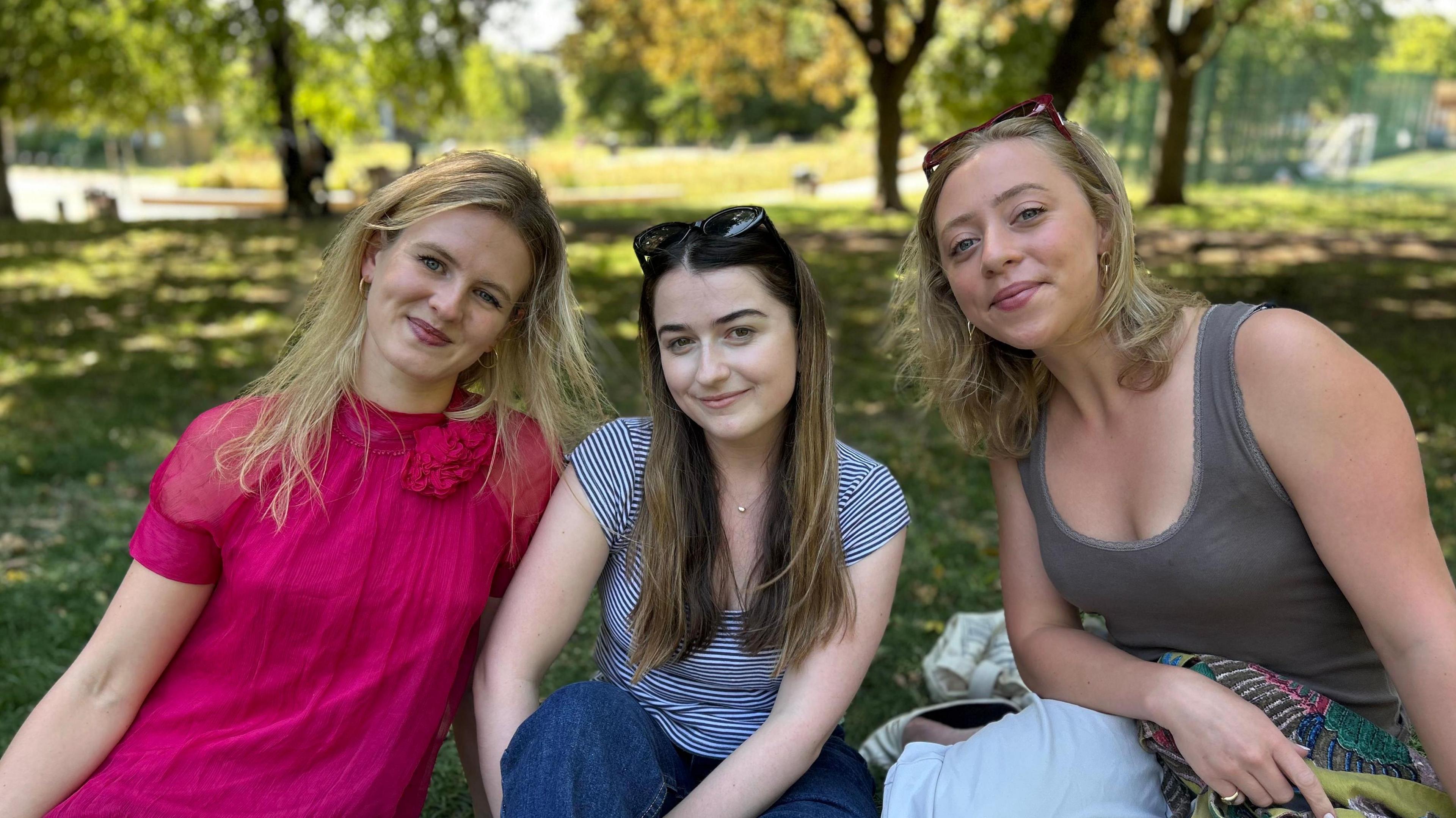 Group of three women sat in park, all smiling at the camera. The woman on left wears a fuchsia pink dress, the woman in the middle, a stripy t-shirt and jeans and then the woman on the right, a sage green vest top. Behind them is a park with leafy trees.