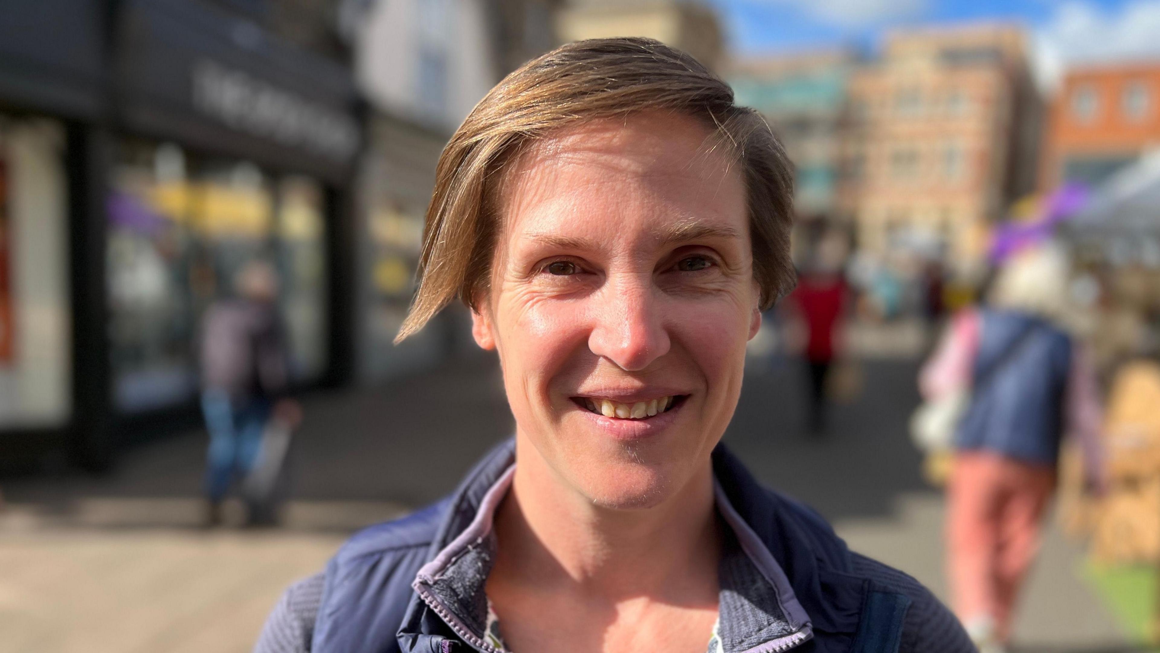 A head and shoulders picture of a woman standing in a pedestrianised street. The background is blurred.