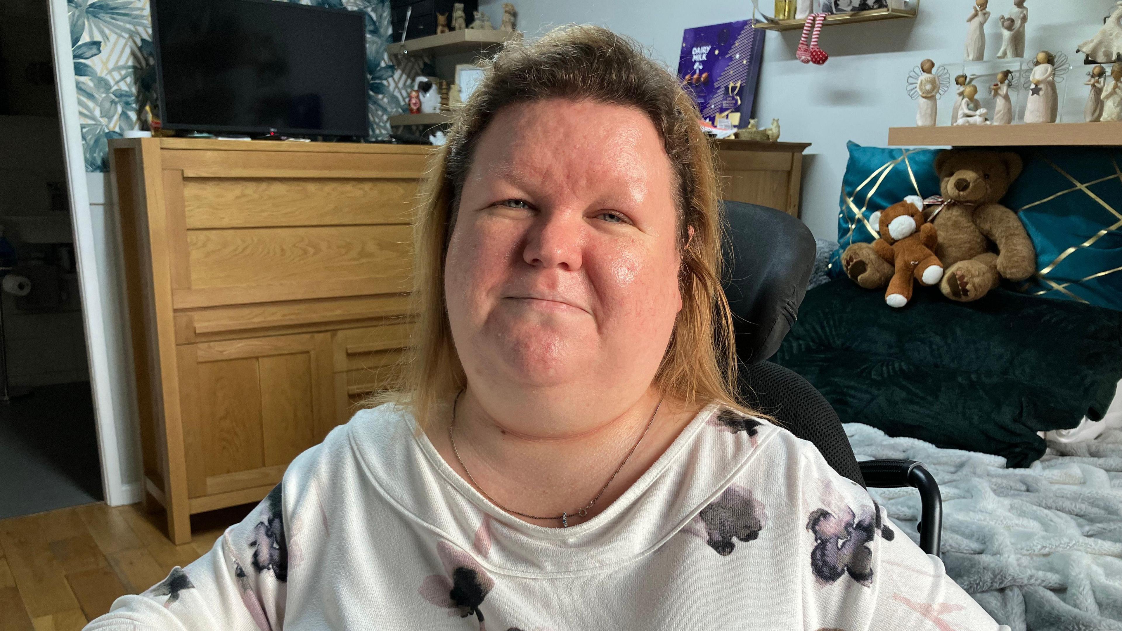 Jennifer Gallacher is wearing a white top with watercolour flowers on it. She has brown hair and is sitting in her bedroom. A bed can be seen behind her with two Teddy bears sitting on a pillow. To the left is a chest of drawers with a TV on top.