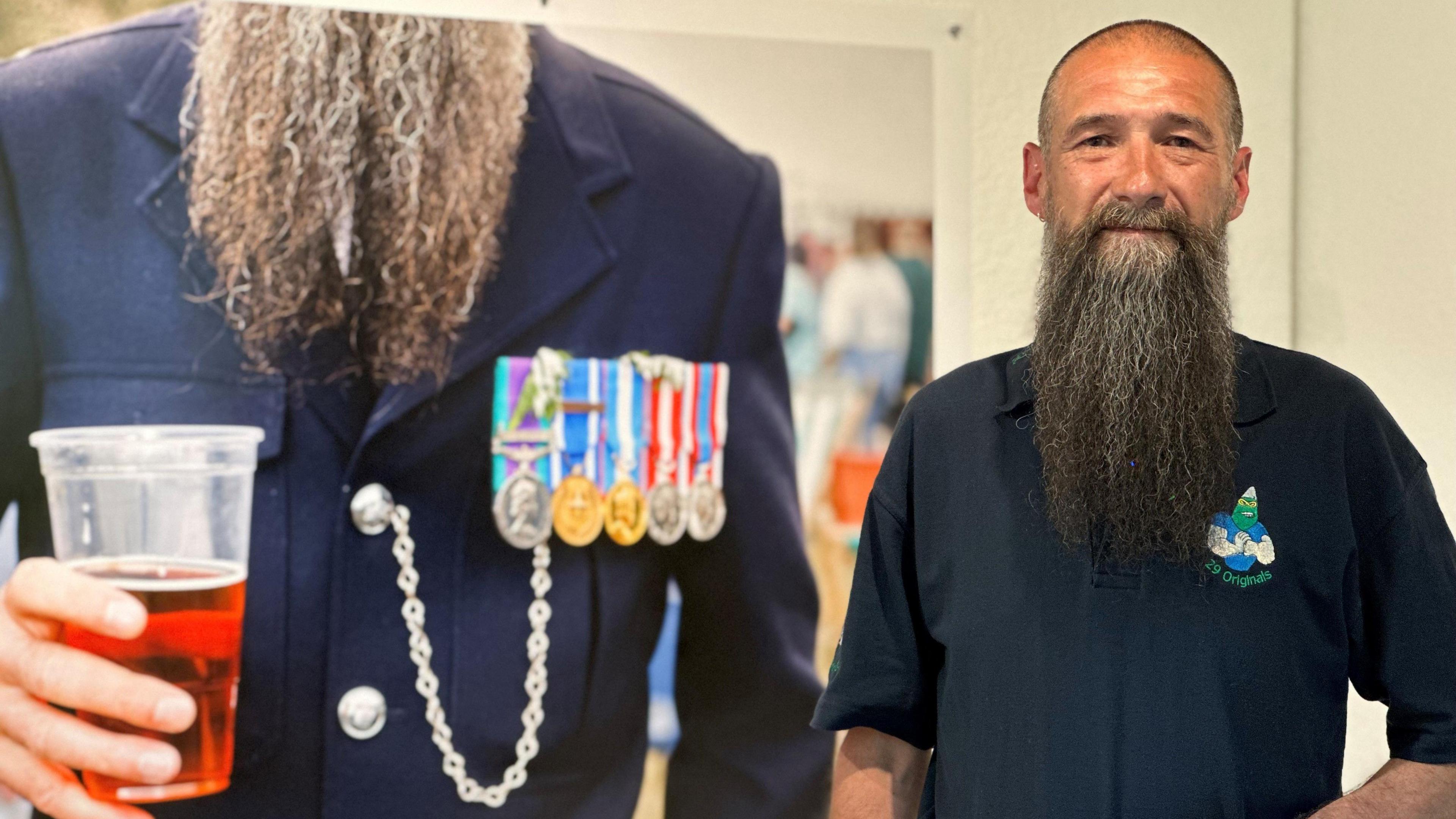 Mark Huston, who has close-cropped dark hair and a long brown beard and is wearing a navy blue T-shirt. He is standing in front of a large print of the photo of himself featured in the exhibition. It shows him from the shoulders down, showing his beard set against his navy blue parade uniform, which has medals and a chain on the left breast. He is holding a plastic pint glass in one hand that is two-thirds full.