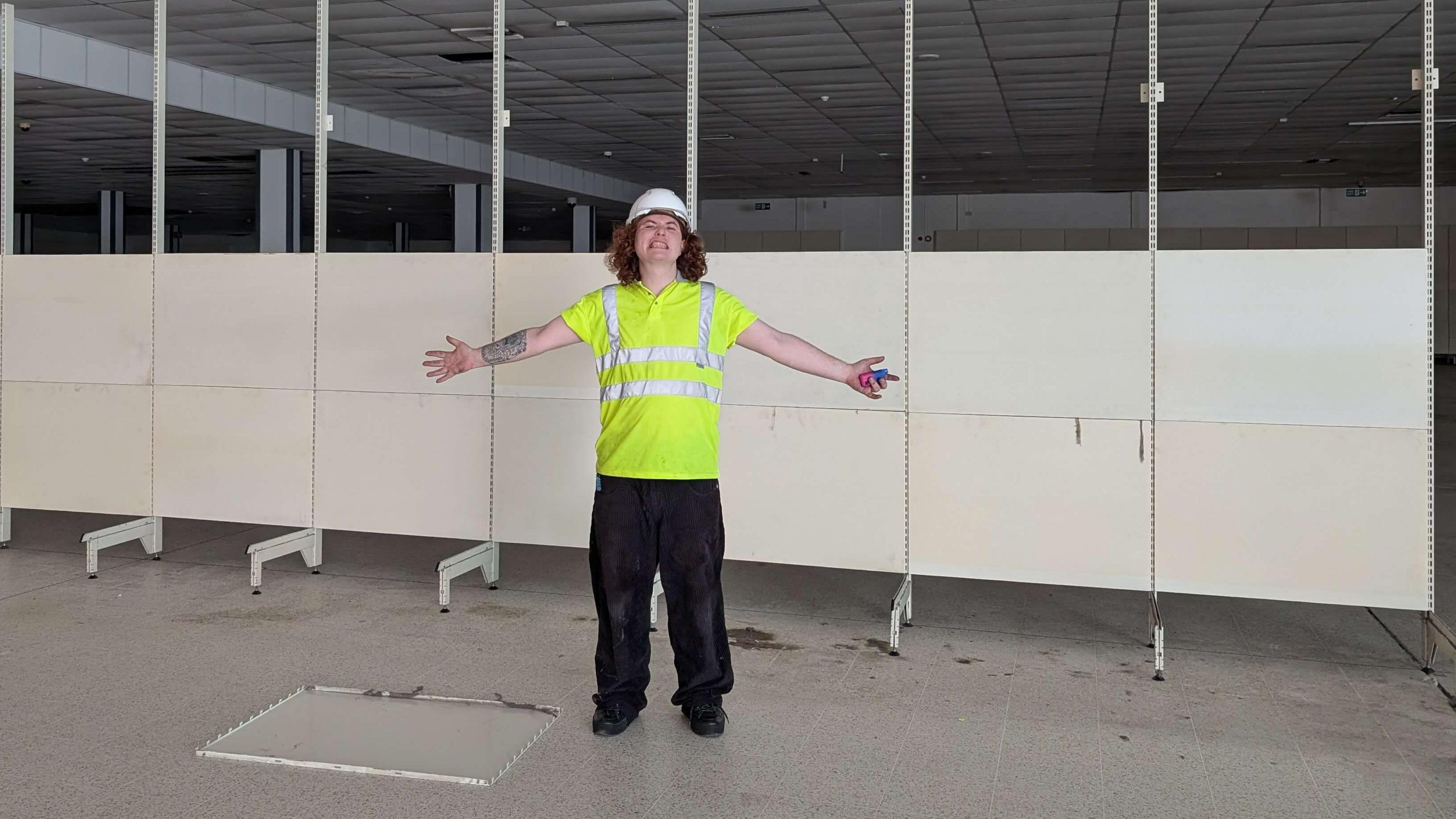 Theodore Godfrey-Cass stands in front of wall partitions which had been put up in the new art gallery before its planned opening. He is wearing a white hard hat and hi-vis jacket.
He has his arms spread wide and he is gurning at the camera.