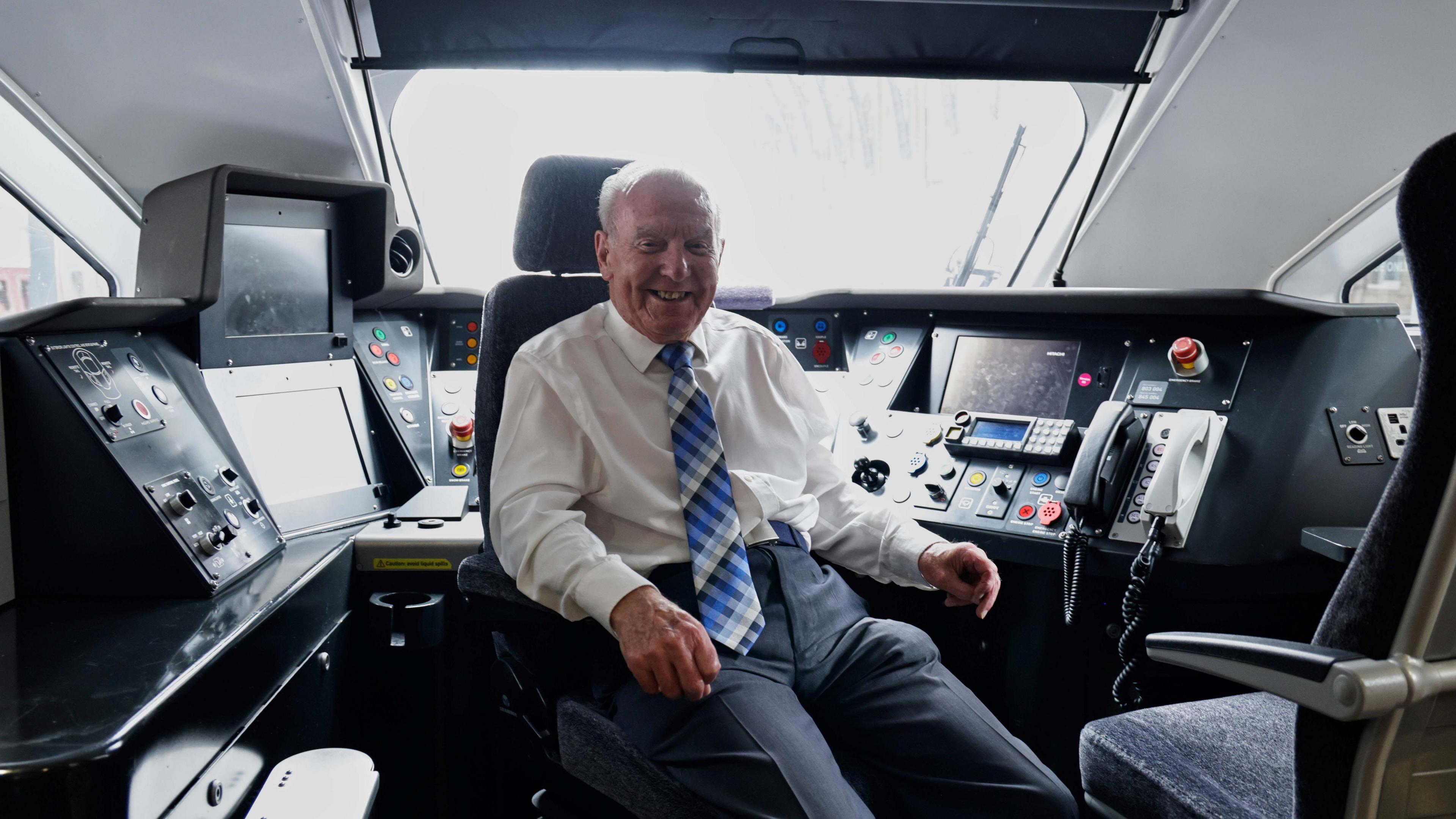 A man with short white hair, a white shirt, a blue patterned tie, dark trousers and a beige and grey jacket sitting in the cab of a train. He is smiling.