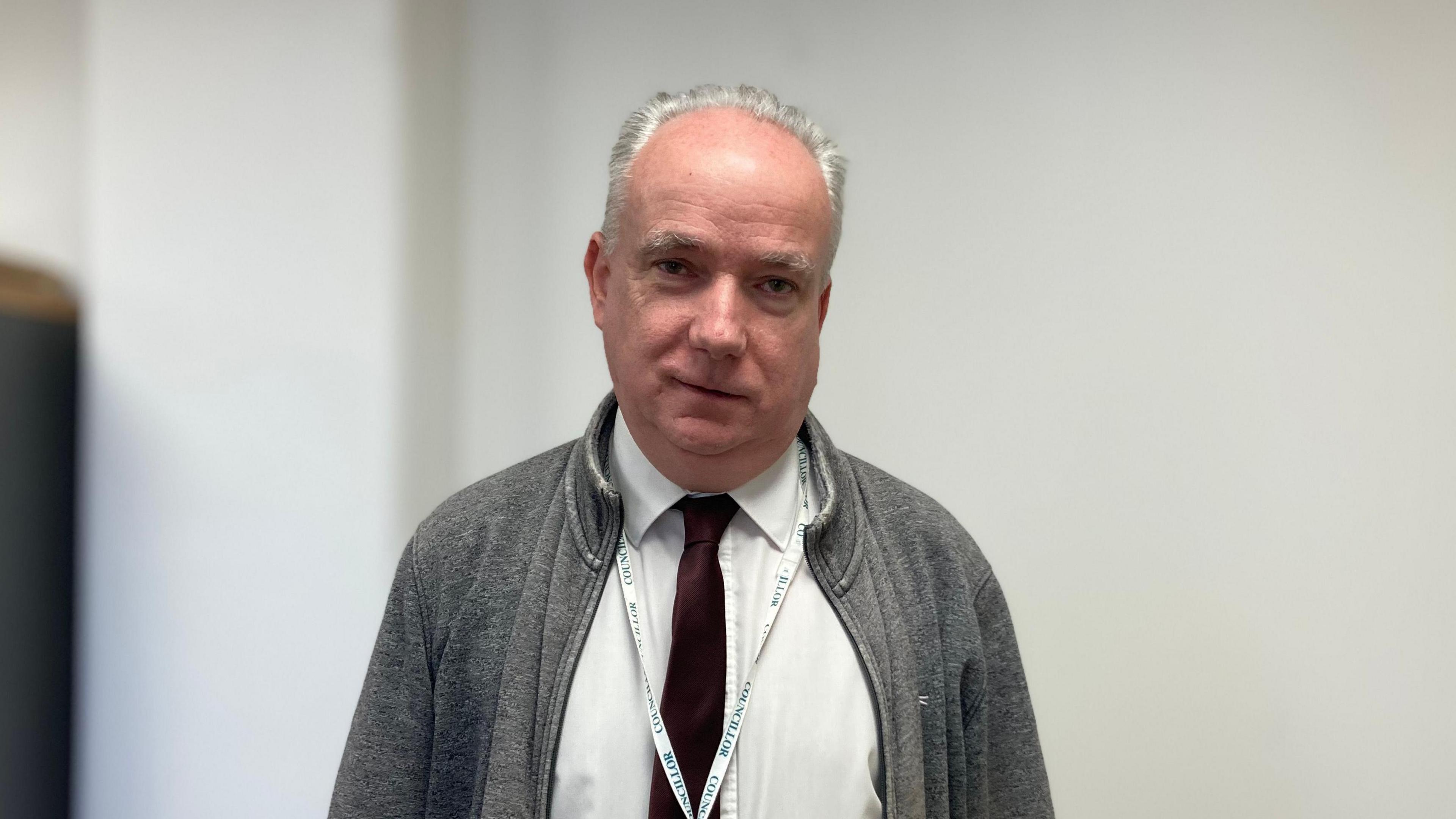 Angus Ellis wearing a grey jacket, white shirt and burgundy tie. He is also wearing a lanyard that says 'councillor'. He is looking at the camera in front of a blurred white background. 