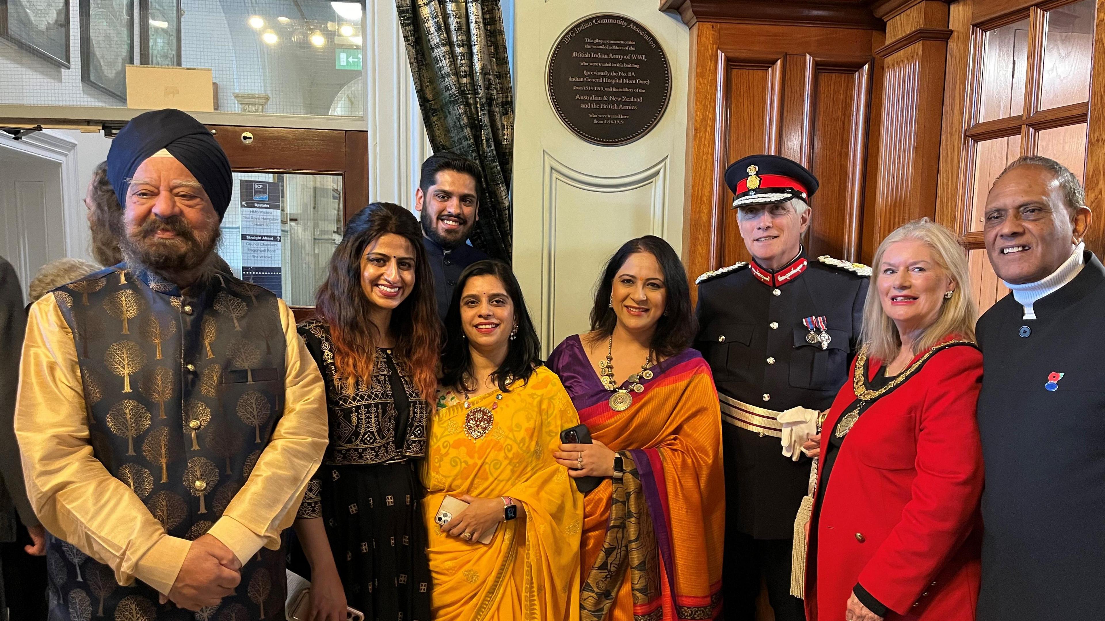 Eight smartly-dressed VIPs stand in front of a wall-mounted plaque inside Bournemouth town hall. Six of the group are from the Indian community.