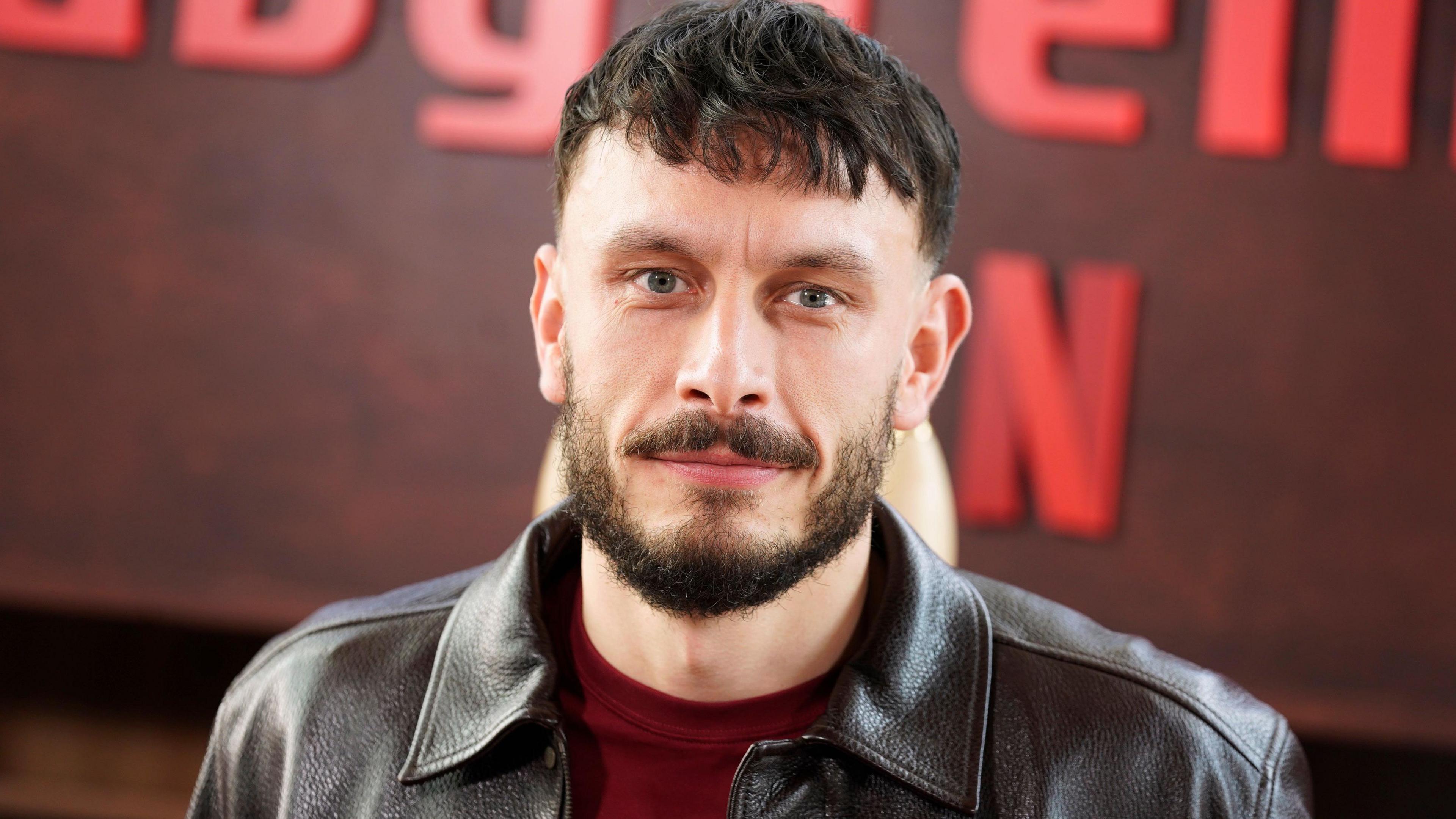 Actor Richard Gadd in a close-up wearing a burgundy sweater and leather jacket, standing in front of a Baby Reindeer sign at the series launch