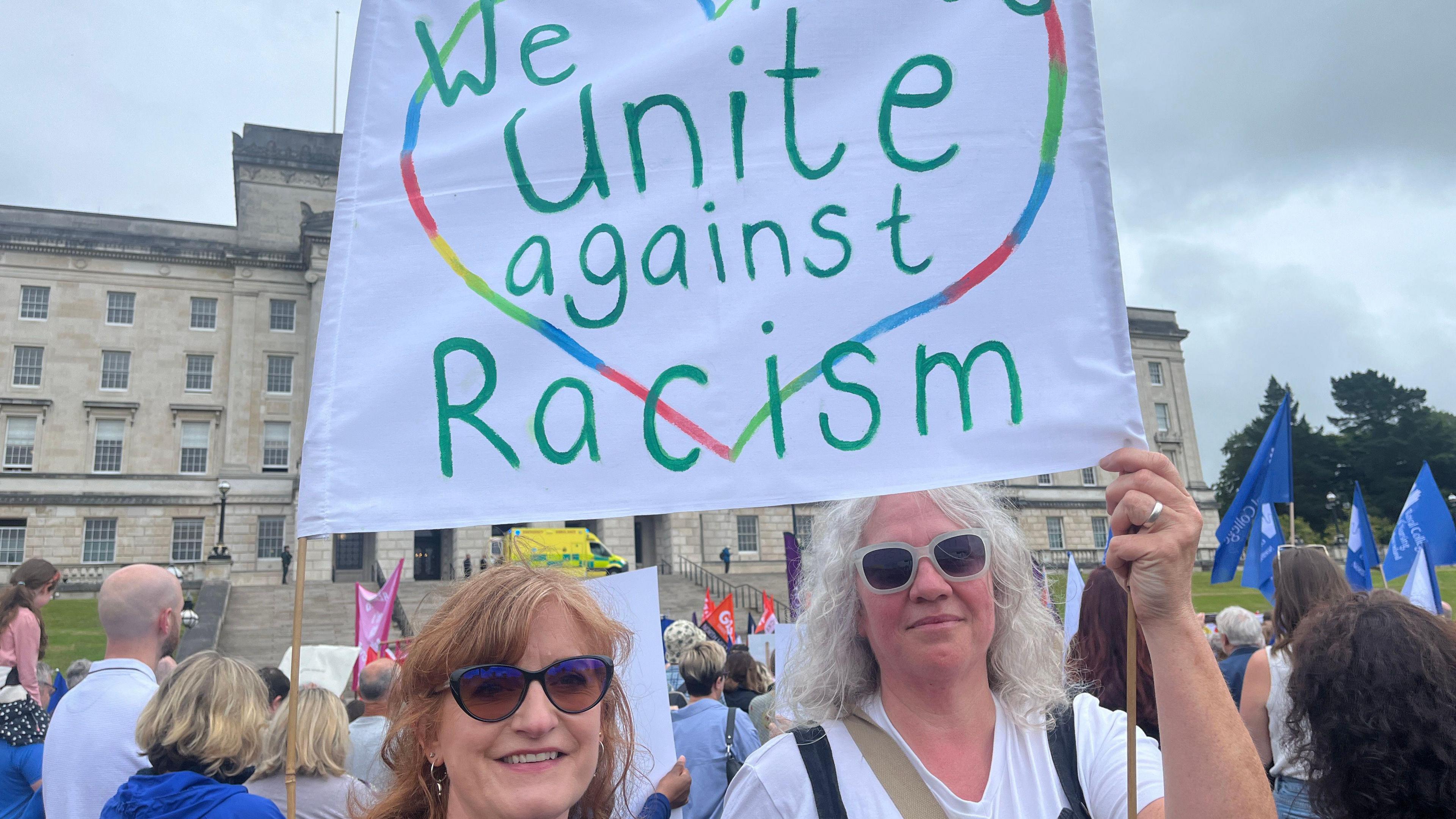 Two women wearing sunglasses holding a banner reading 'We unite against Racism' in front of Stormont