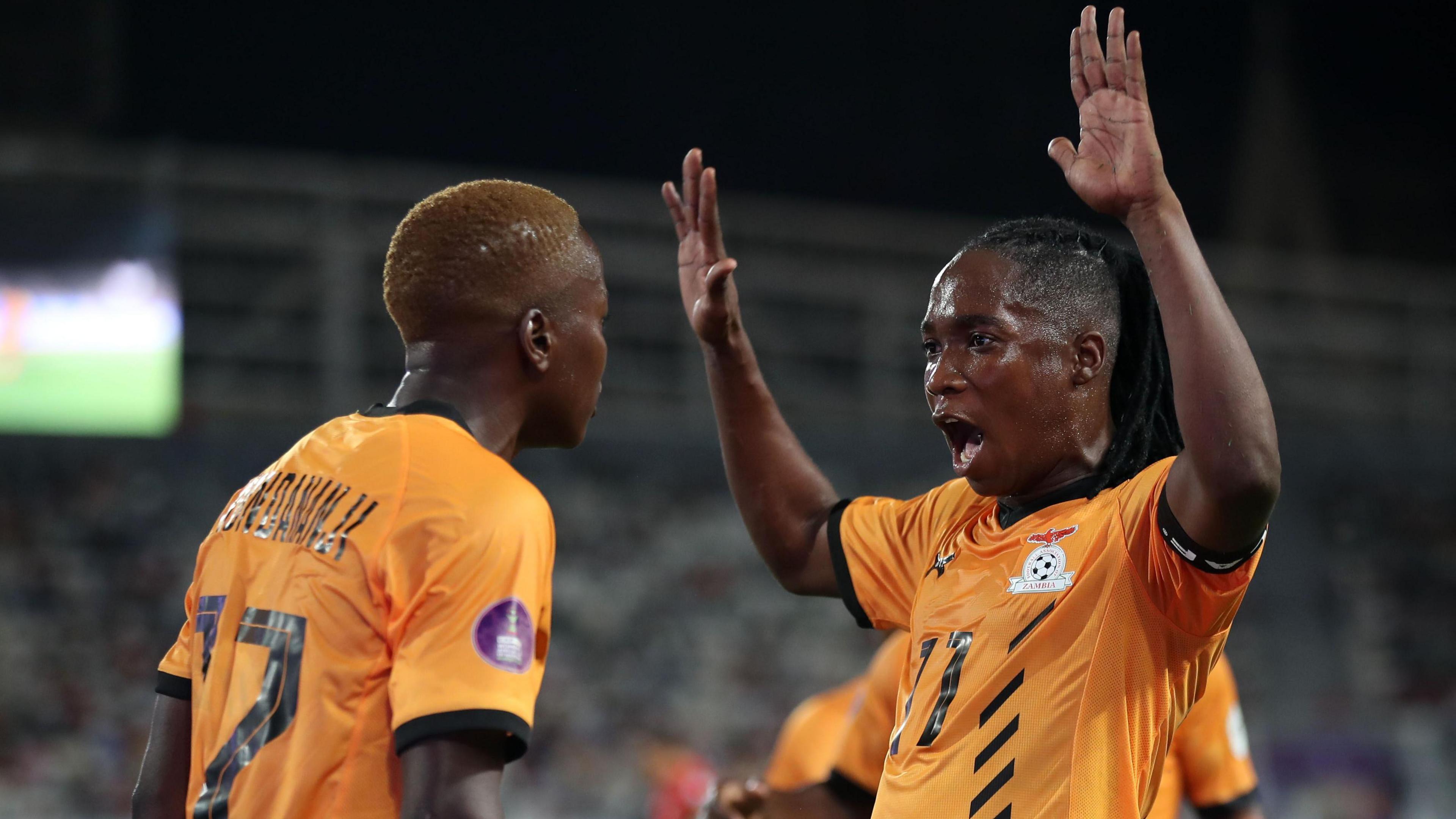 Racheal Kundananji and Barbra Banda, both in orange Zambia kit, are seen from waist up celebrating, with Banda shouting and raising both her arms above her head with palms open