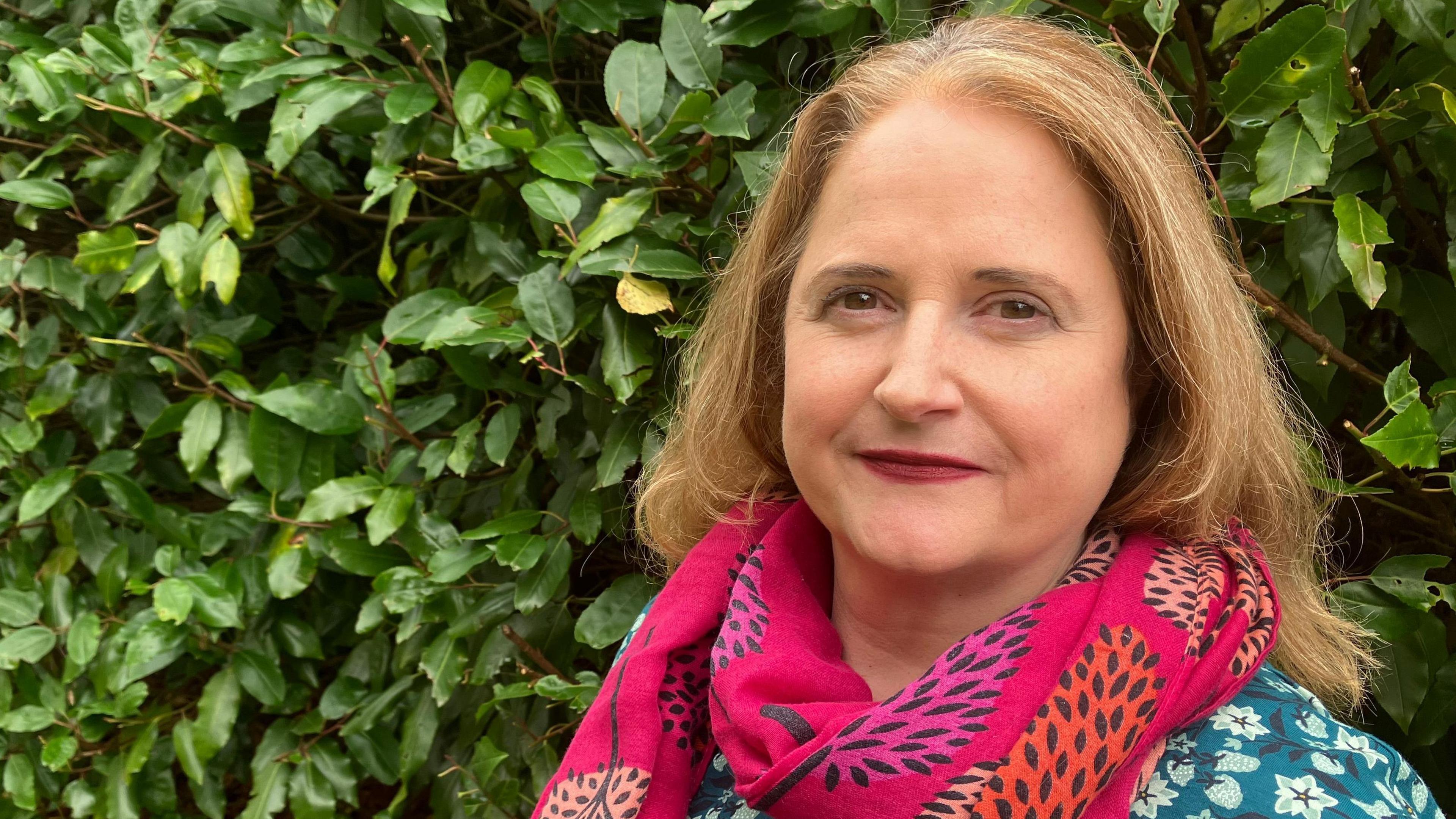 Lara Pringle looks directly at the camera. She has light brown hair and is wearing a pink scarf with images of trees on it, and a green blouse with flowers on it. Behind her is a hedge.