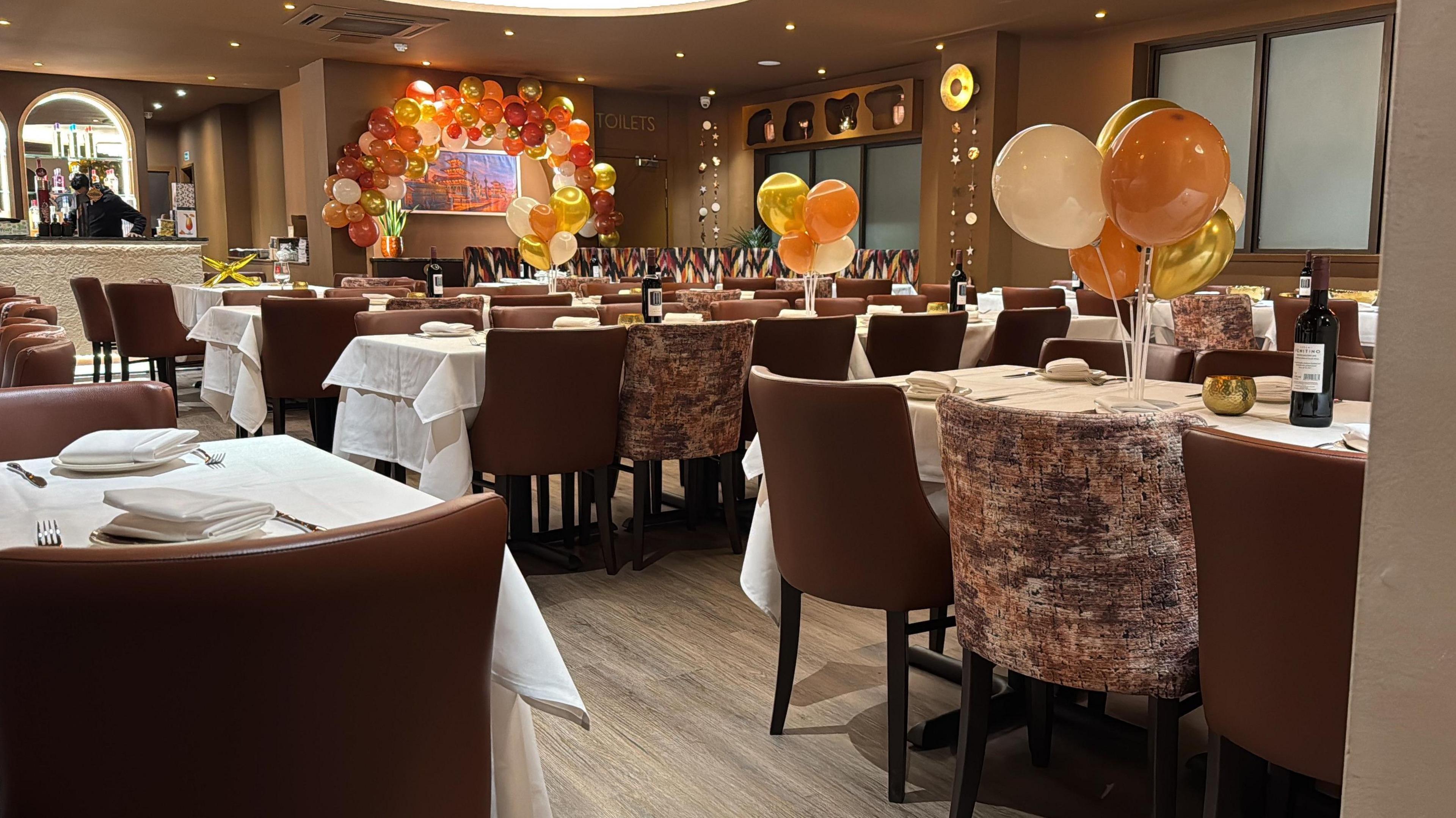 A restaurant with taupe walls, brown leather-style and velvet chairs, with gold balloon displays distributed around the room.