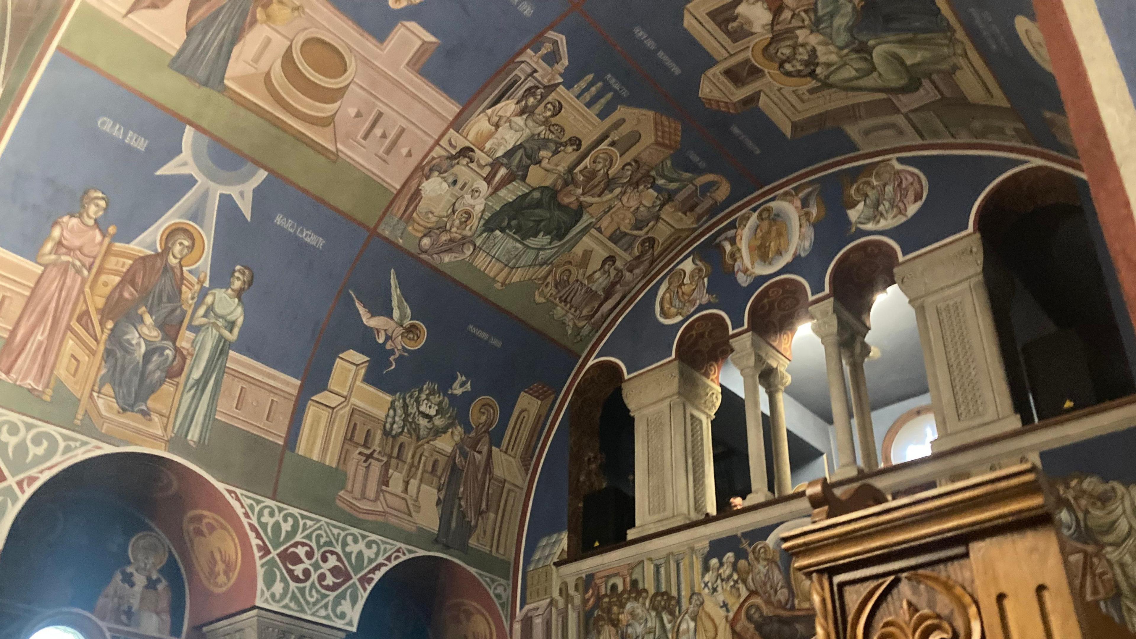 Frescos showing religious paintings in different colours which are mainly blue, brown, green and white. The ceiling is arched and there are cream pillars at the top.