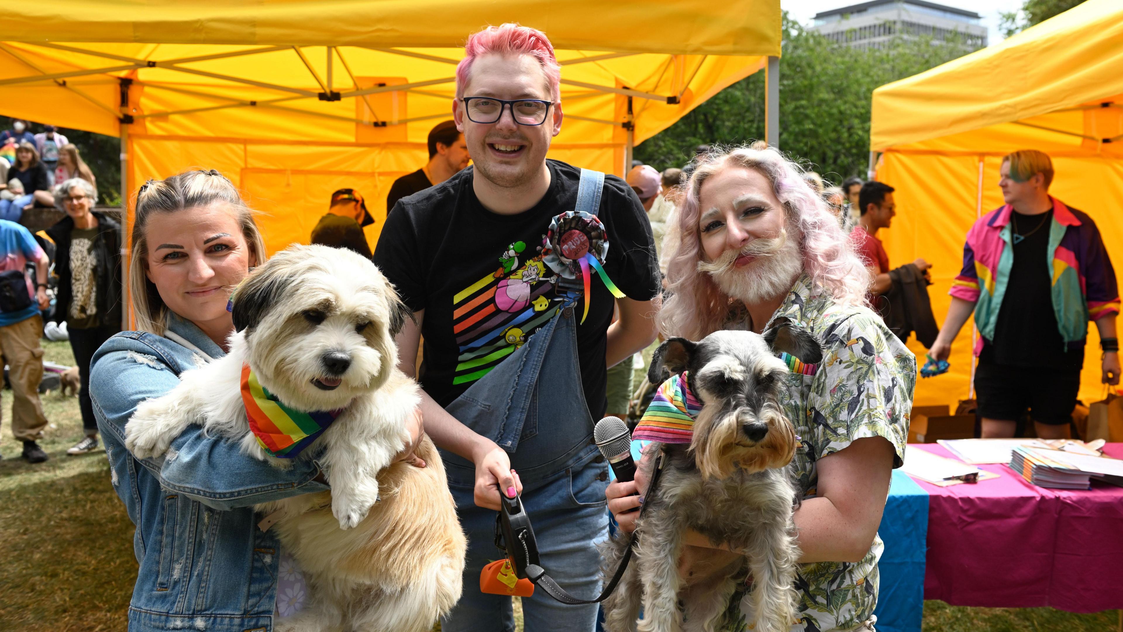 Three people smiling at the camera, two of them, either side, are holding dogs wearing pride handkerchiefs 